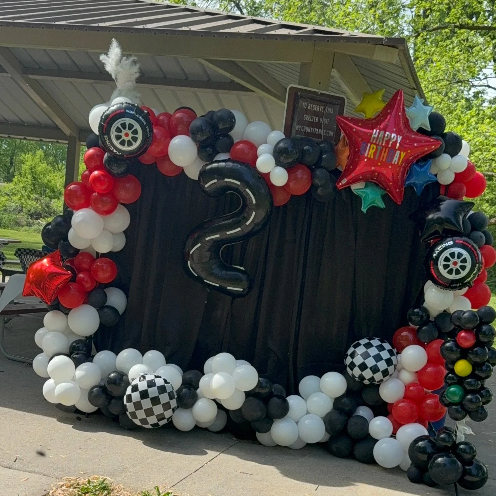 Close-up of a smiling woman with curly hair, wearing glasses, hoop earrings, and layered necklaces, indoors.