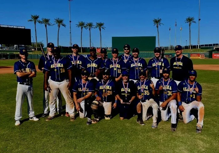 A team of young baseball players in blue uniforms with medals around their necks, posing on a baseball field at a stadium with trees and scoreboard in the background.