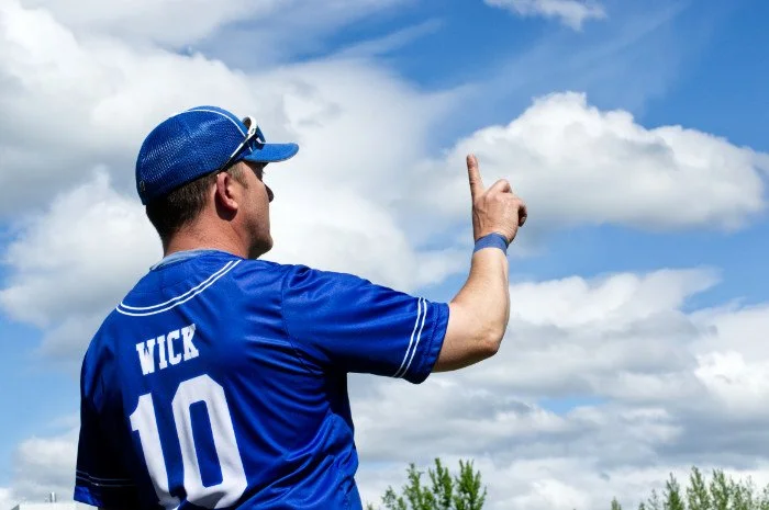 A man wearing a blue baseball jersey with the name WICK and the number 10, a blue cap, and sunglasses, pointing upwards at the sky.