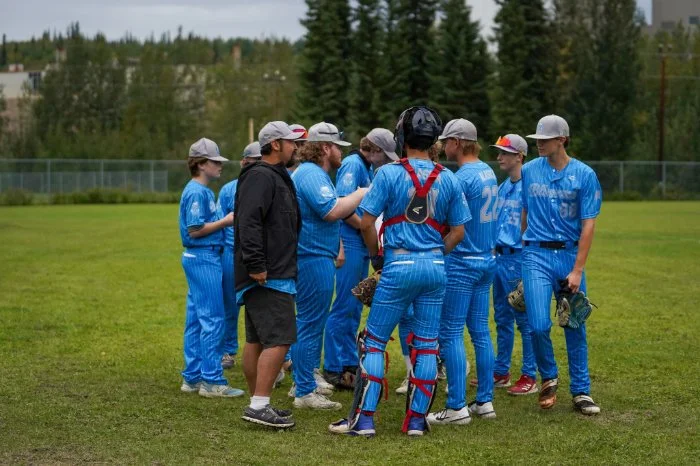 A youth baseball team in blue uniforms gathers in a huddle on a grassy field during daytime.