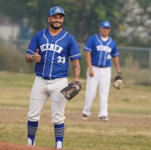 Two baseball players in blue uniforms on a field, one smiling and giving a thumbs up, the other standing in the background.