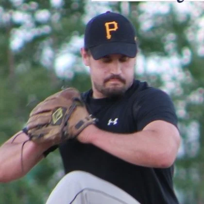 Male baseball player wearing a black cap with a yellow P, black T-shirt, and brown glove, in a baseball field.