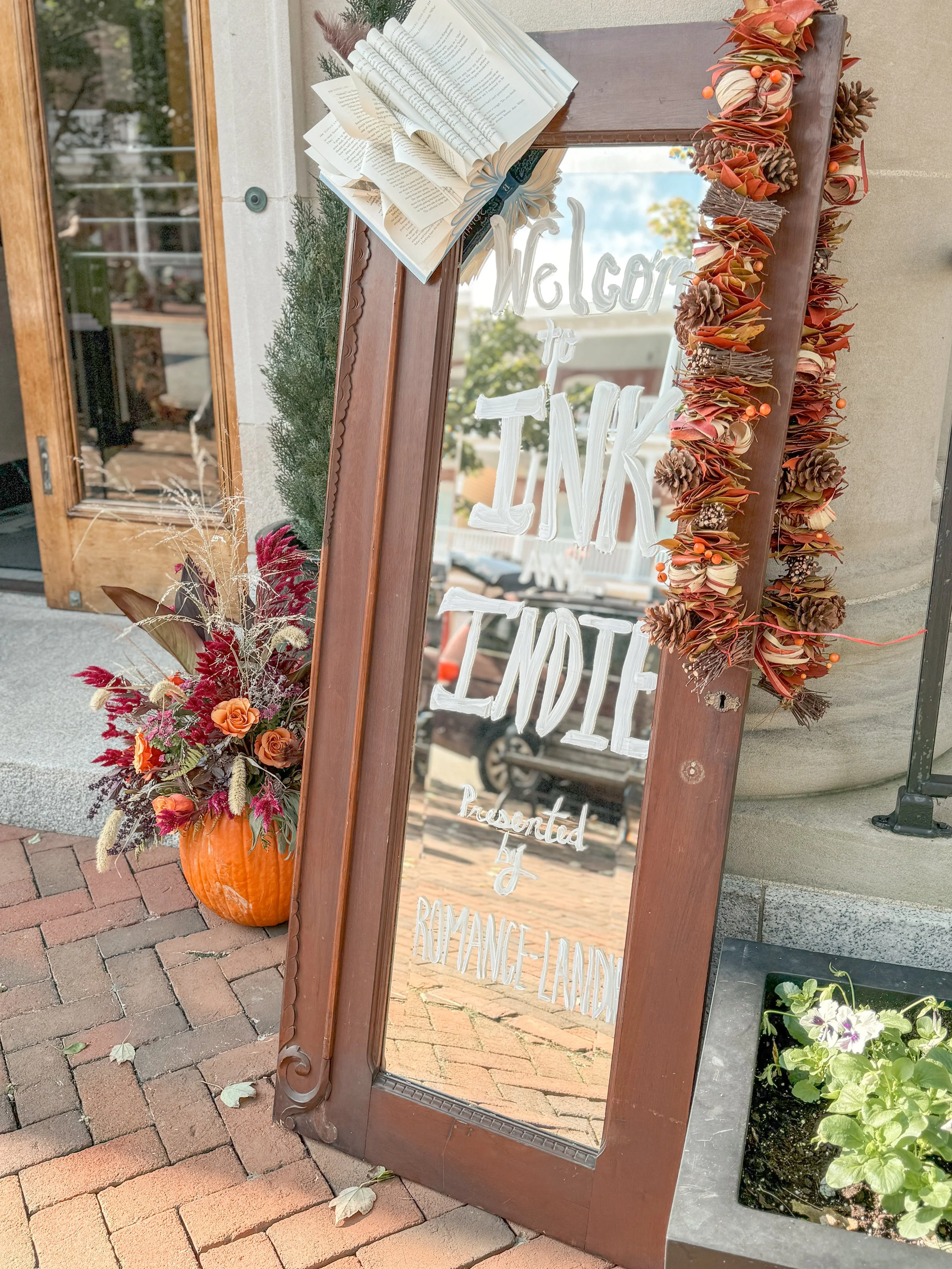 Decorative outdoor sign with a mirror reading 'Welcome to JNK Indie,' surrounded by fall-themed decorations including a pumpkin filled with flowers and foliage, a garland of pinecones and leaves, and a potted plant with purple flowers.