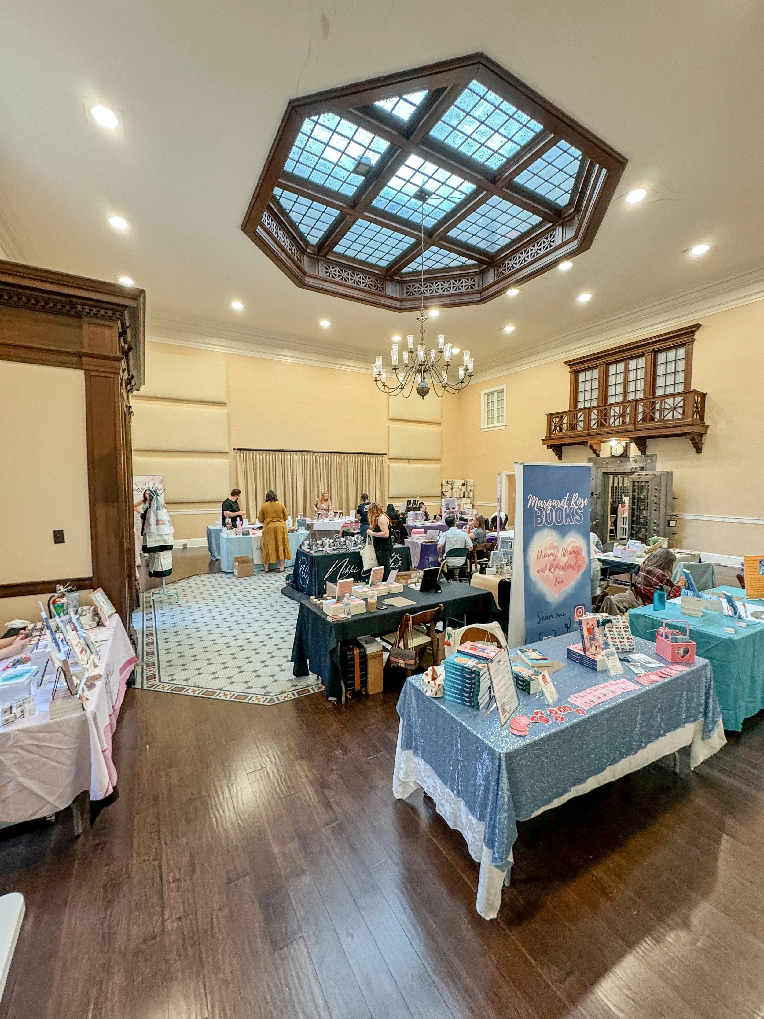 An indoor event space with tables displaying books and merchandise, with some people browsing and engaging with vendors.