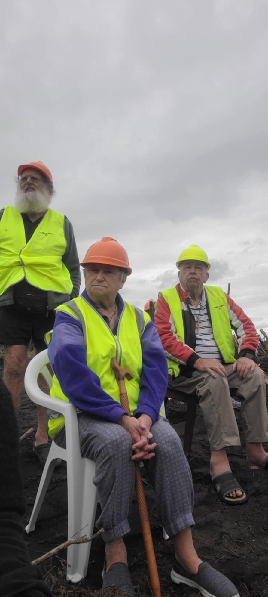 Three elderly people wearing neon safety vests and hard hats sitting and standing outdoors under a cloudy sky.