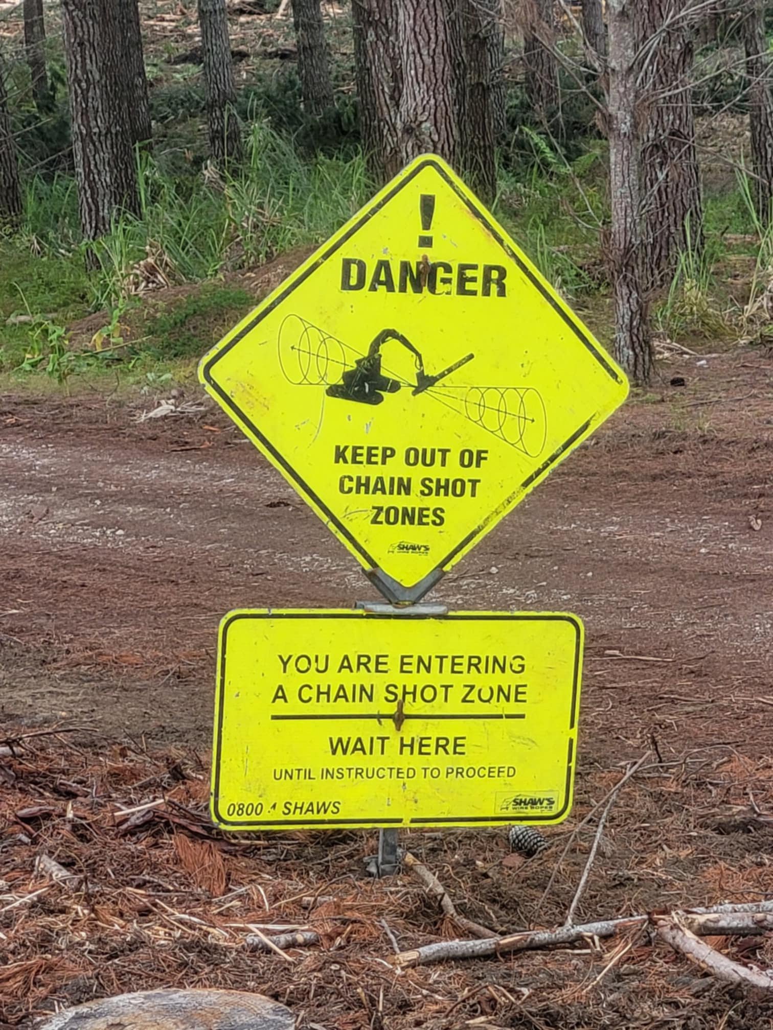 Warning sign in a forest area cautioning about chain shot zones. The top sign reads “Danger: Keep Out of Chain Shot Zones” with an exclamation mark and an image of machinery. Below it, another sign states, “You Are Entering a Chain Shot Zone. Wait He