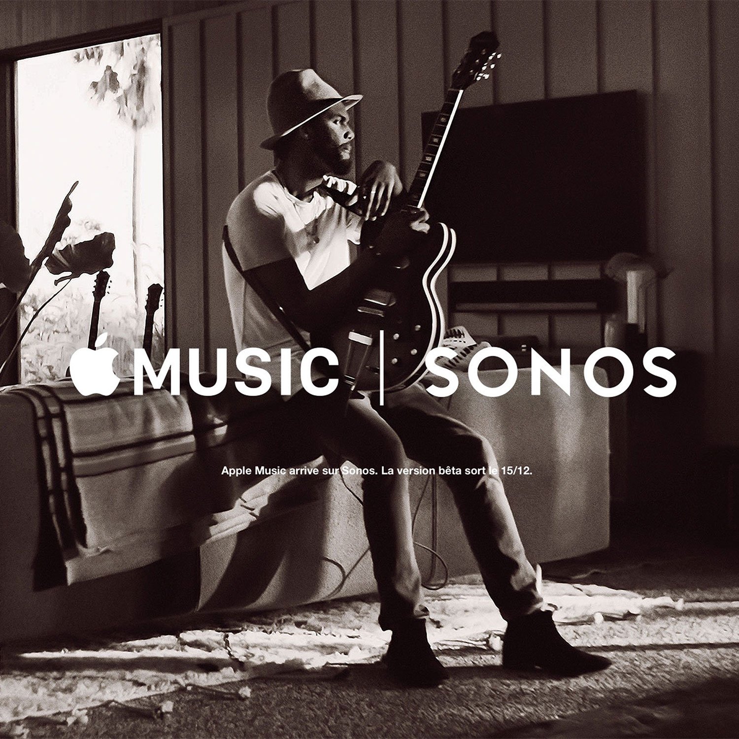 Black and white photo of a man wearing a fedora hat, sitting on a couch, holding a guitar. There is a TV on the wall behind him and some plants nearby. The image features the Apple Music and Sonos logos, with text announcing the arrival of Apple Music on Sonos