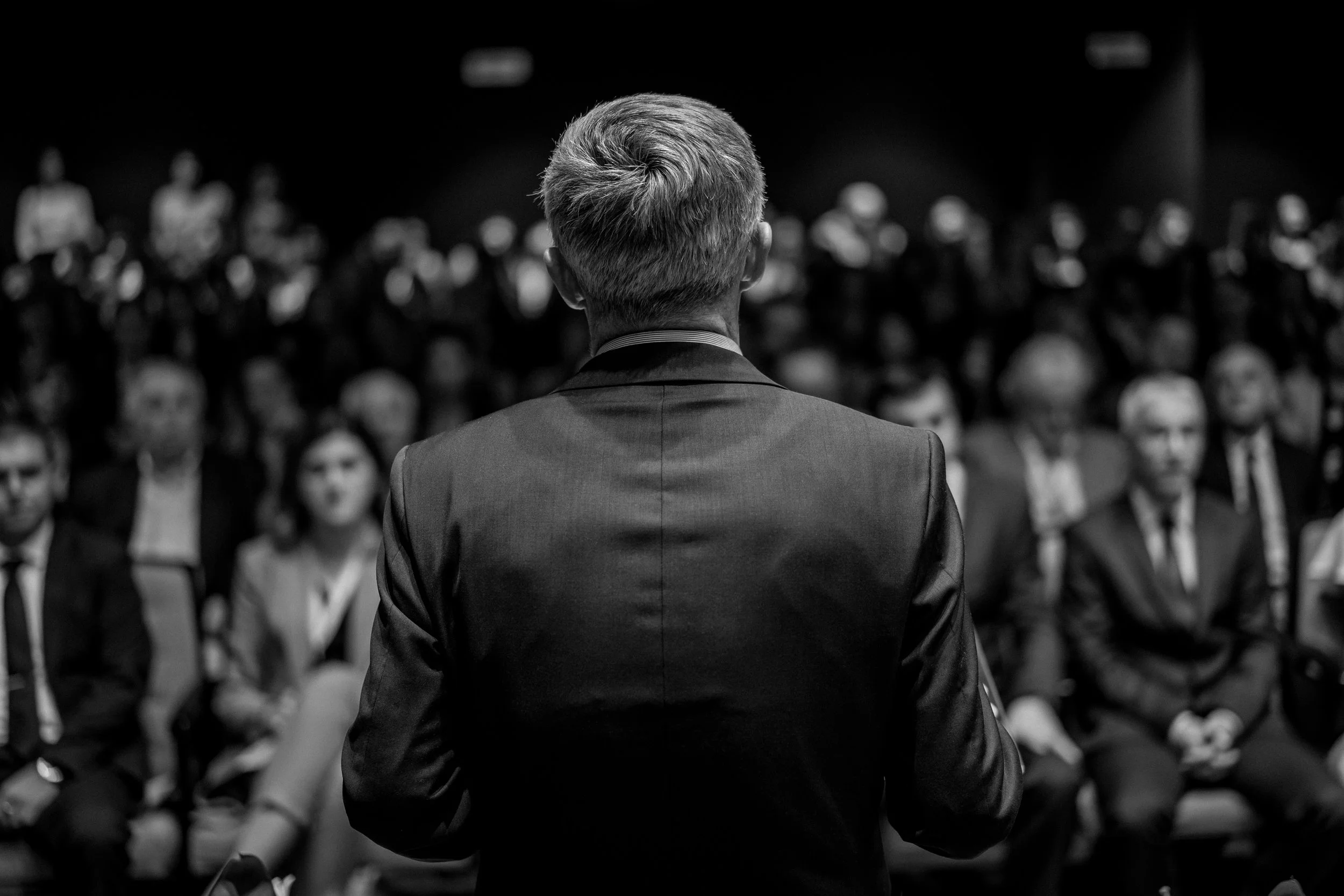 Rear view of a senior official addressing a seated audience in a formal policy briefing or conference setting