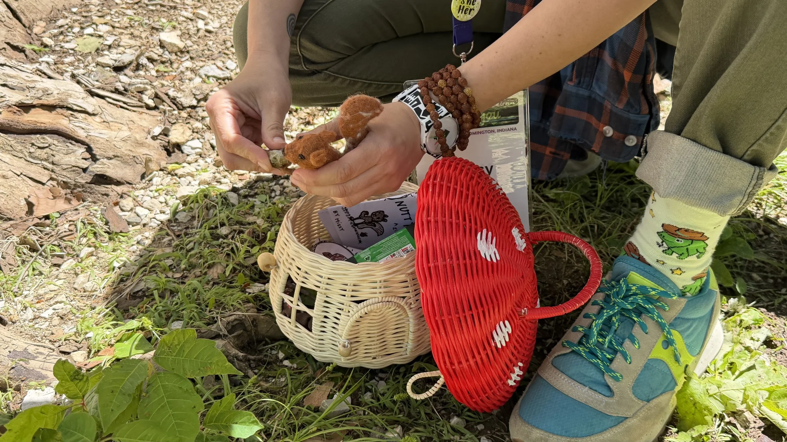 Person crouching outdoors on a gravelly path, handling a small rock. They are wearing colorful shoes, striped socks with a dinosaur design, and have a beaded bracelet. A red woven basket and a paper bag with snacks are on the ground nearby.