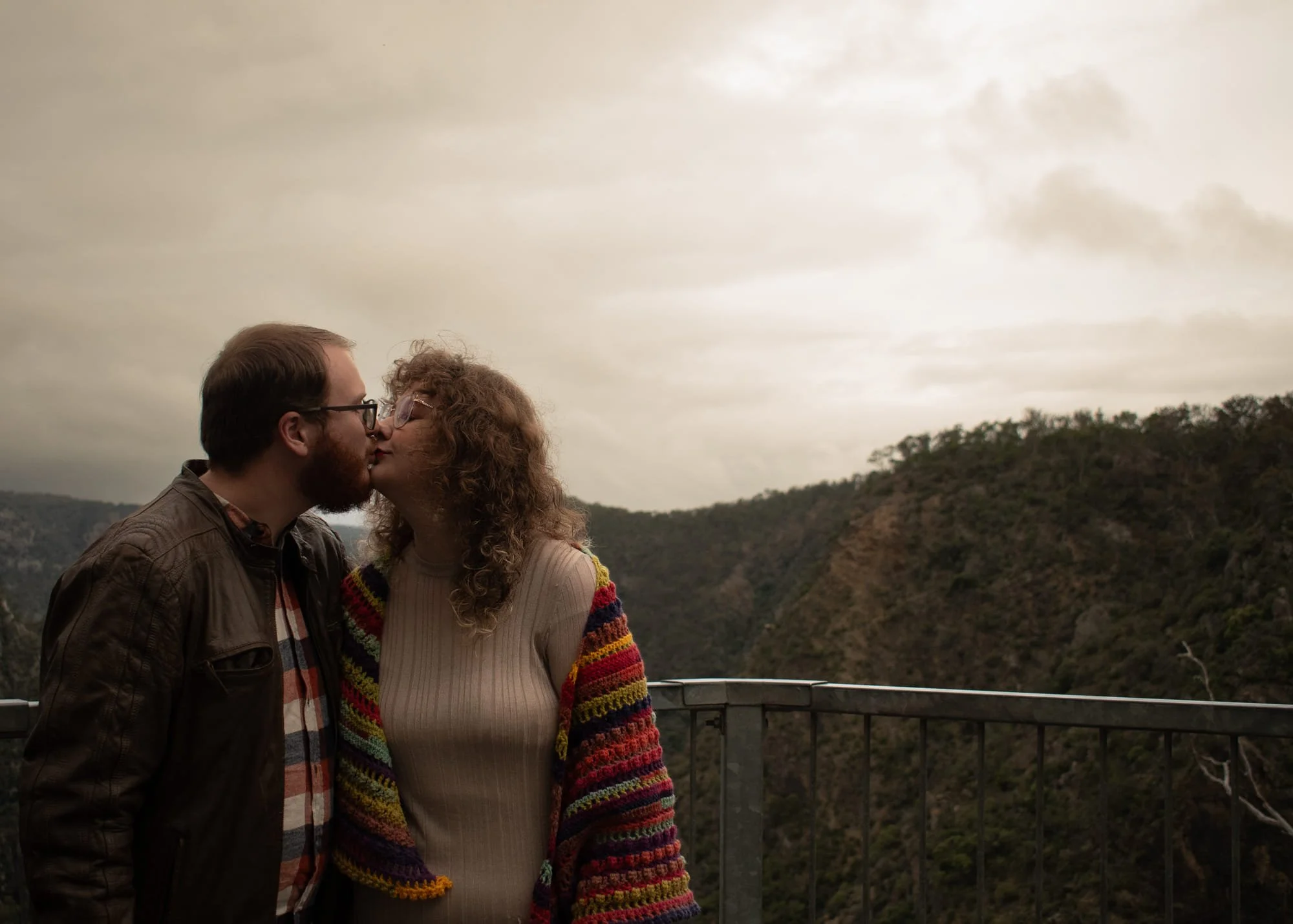 A couple sharing a kiss on a balcony overlooking a hilly landscape under a cloudy sky.