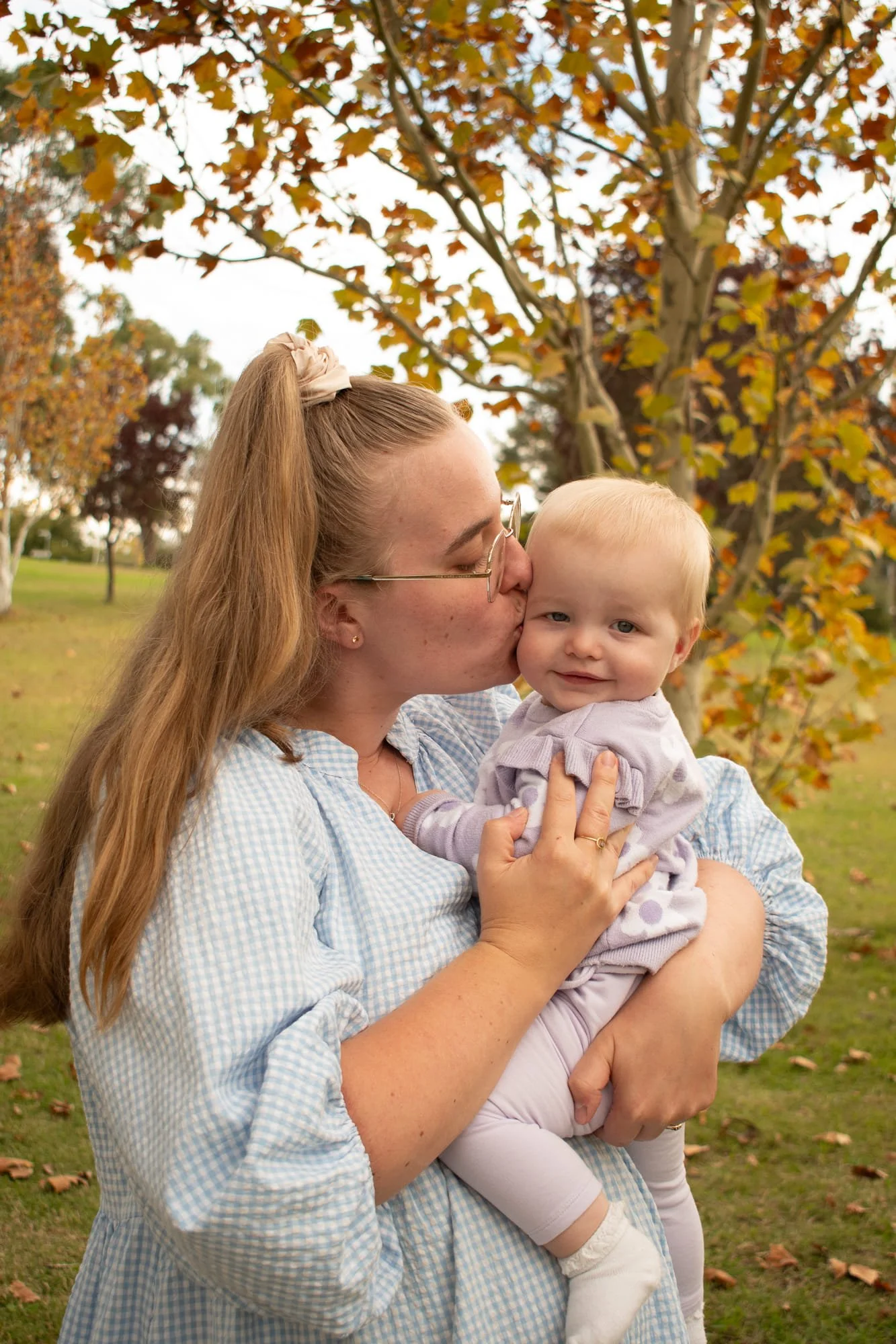 A woman with long red hair in a ponytail, wearing glasses and a light blue checkered shirt, kisses a blonde toddler girl on the cheek while holding her in an outdoor park with a tree and fallen autumn leaves in the background.