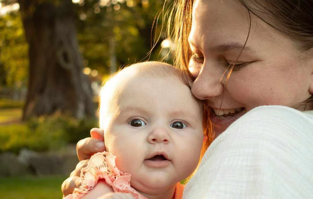A woman holding a baby outdoors during sunset, with trees and grass in the background.