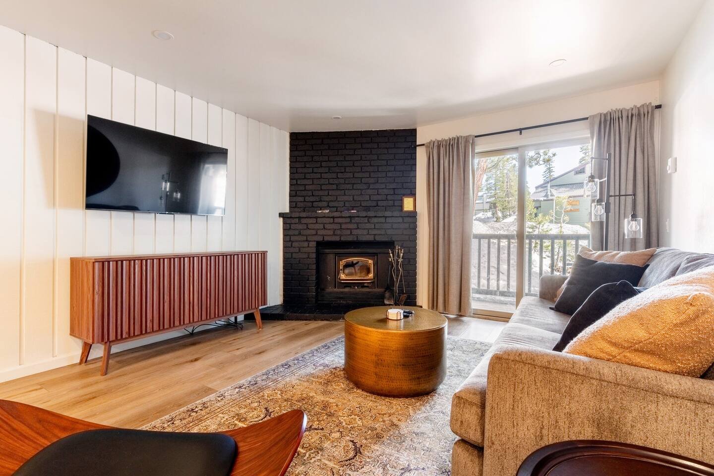 Living room with a beige sofa and black and gold pillows, a round wooden coffee table, a black brick fireplace, a flat-screen TV on a white-paneled wall, a wooden sideboard, and sliding glass doors with curtains leading to an outdoor deck.