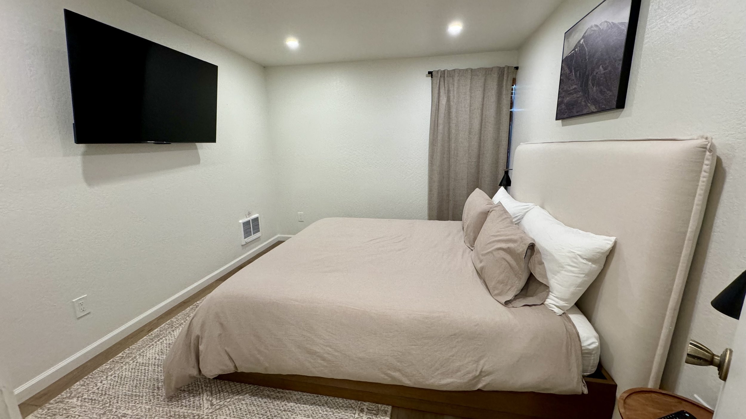 A bedroom with a beige upholstered bed, beige bedding, and pillows. There is a wall-mounted flat-screen TV on the left wall, a framed picture on the right wall, a window with beige curtains, and a textured carpet.