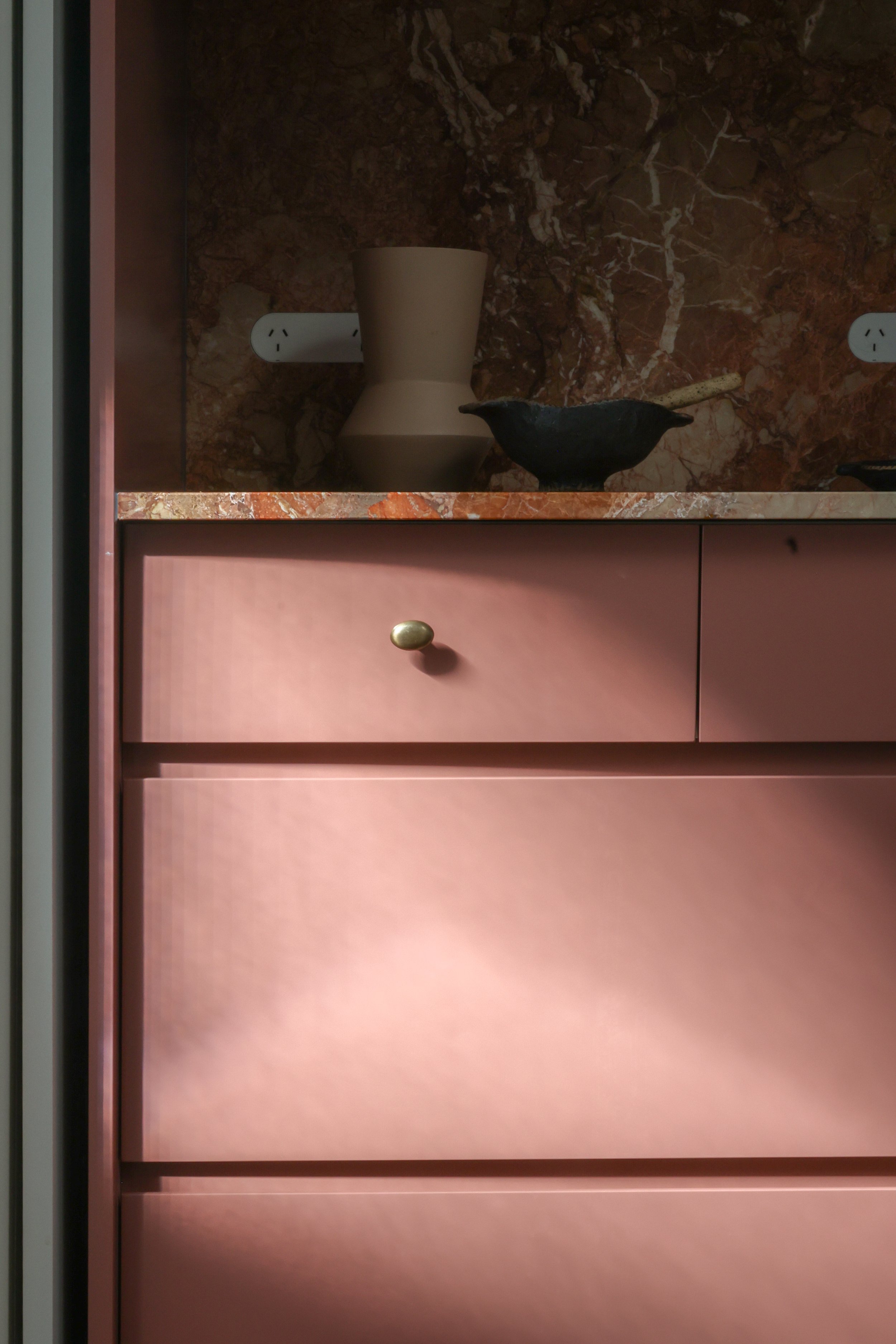 A pink appliance cupboard with a gold knob, a marble countertop, and decorative items including a beige vase and a black bird-shaped object, set against a marble backsplash.