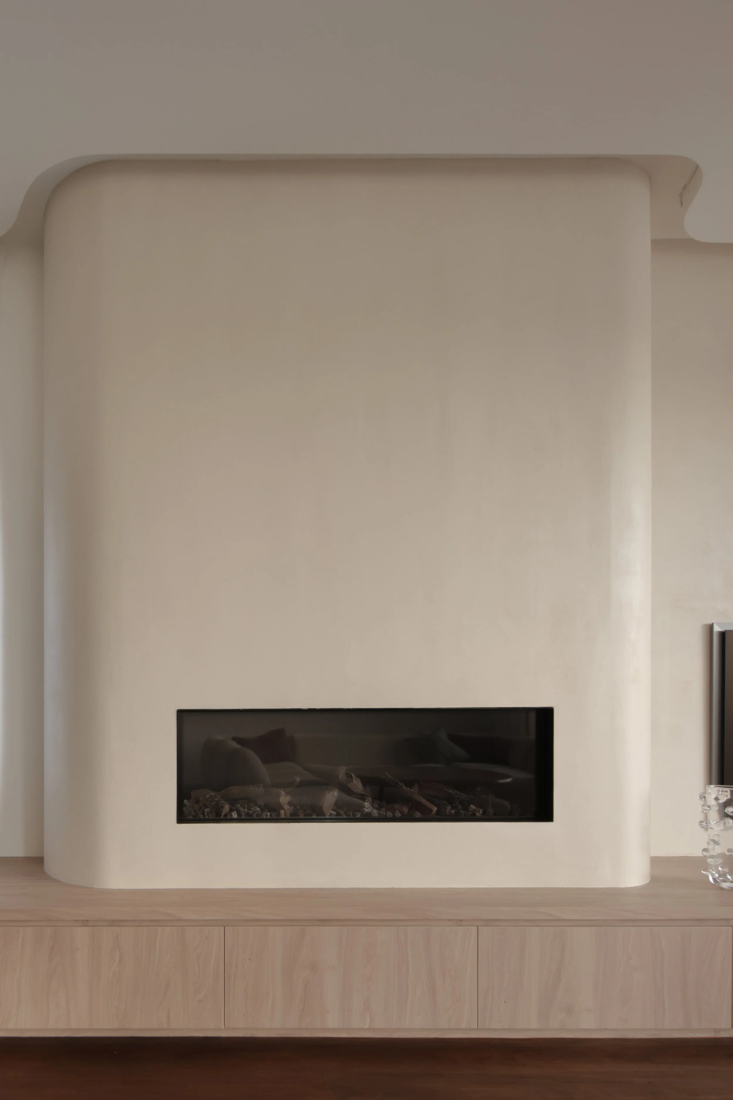 Modern white fireplace with glass front, built into a rounded wall, situated above a light wooden cabinet in a room with wood flooring.