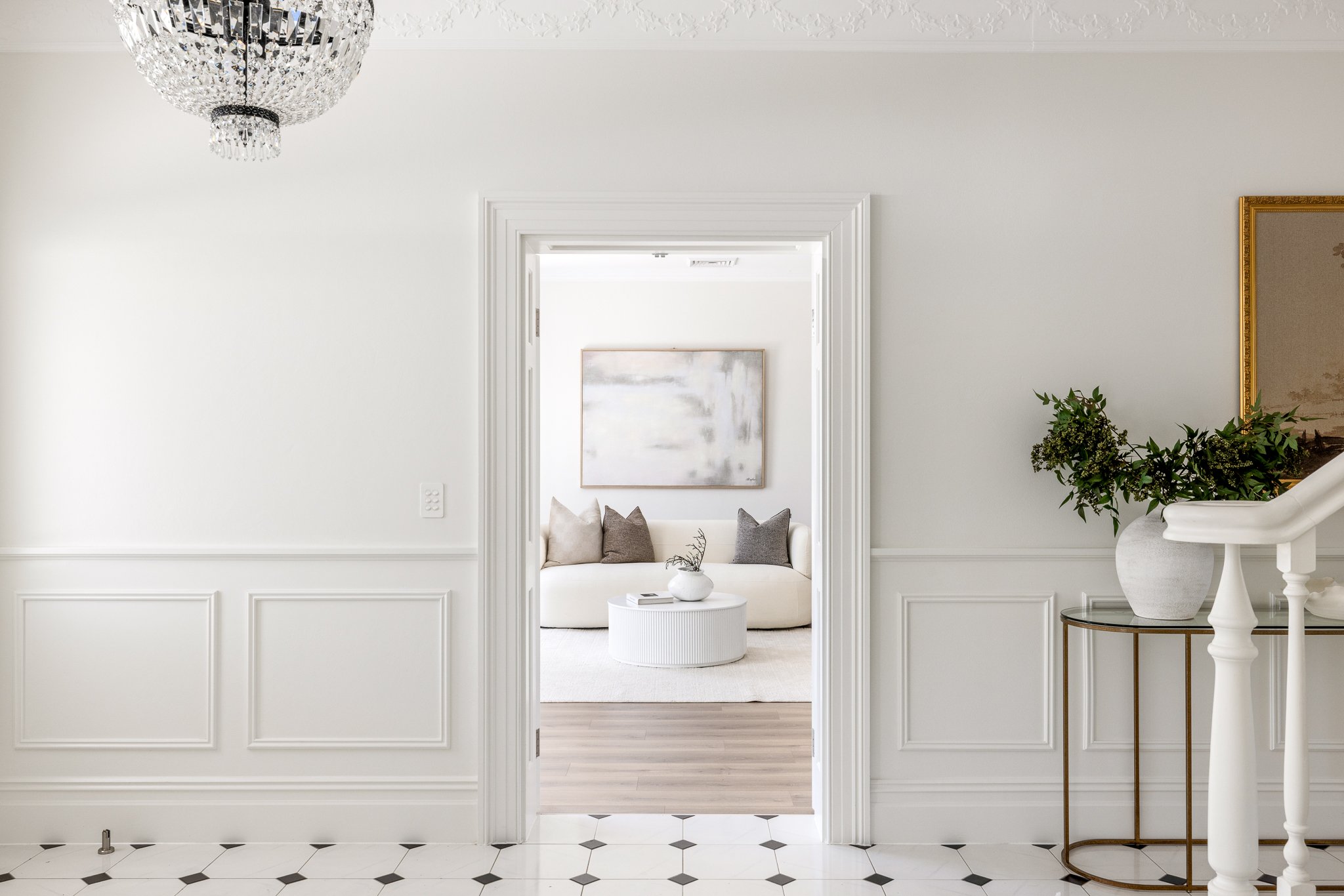 A white marble tiled floor leading to a living room with white walls and a white sofa with three gray pillows. A large abstract painting is above the sofa. To the right, a gold-framed mirror and a white pot with greenery sit on a small glass-topped table. A crystal chandelier hangs from the ceiling.