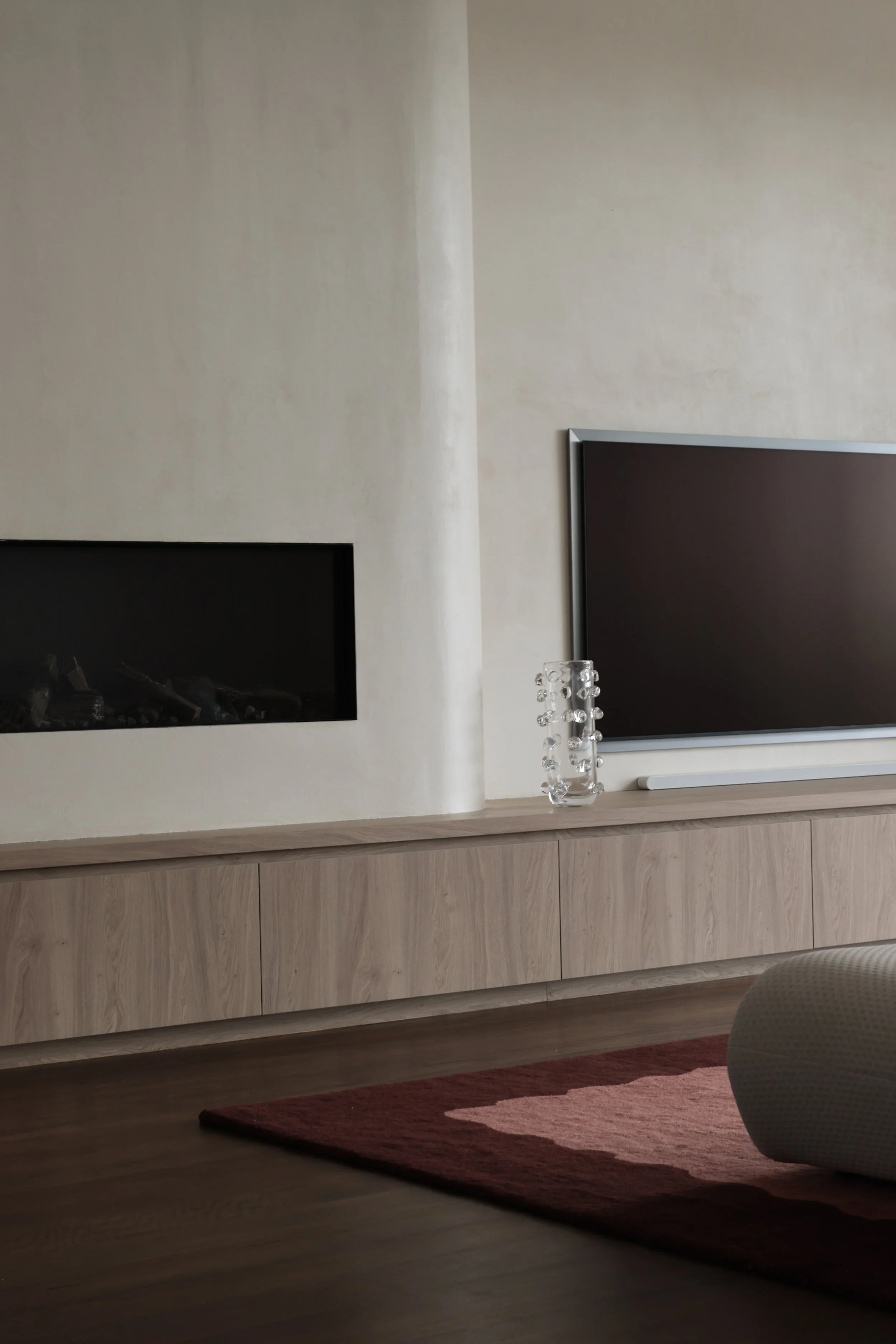 Living room with a large curved fireplace, modern flat-screen TV mounted on the beige wall, a built-in wooden cabinet underneath, a clear glass vase with glass spheres on top of the cabinet, and a bicoloured area rug on the wooden floor.