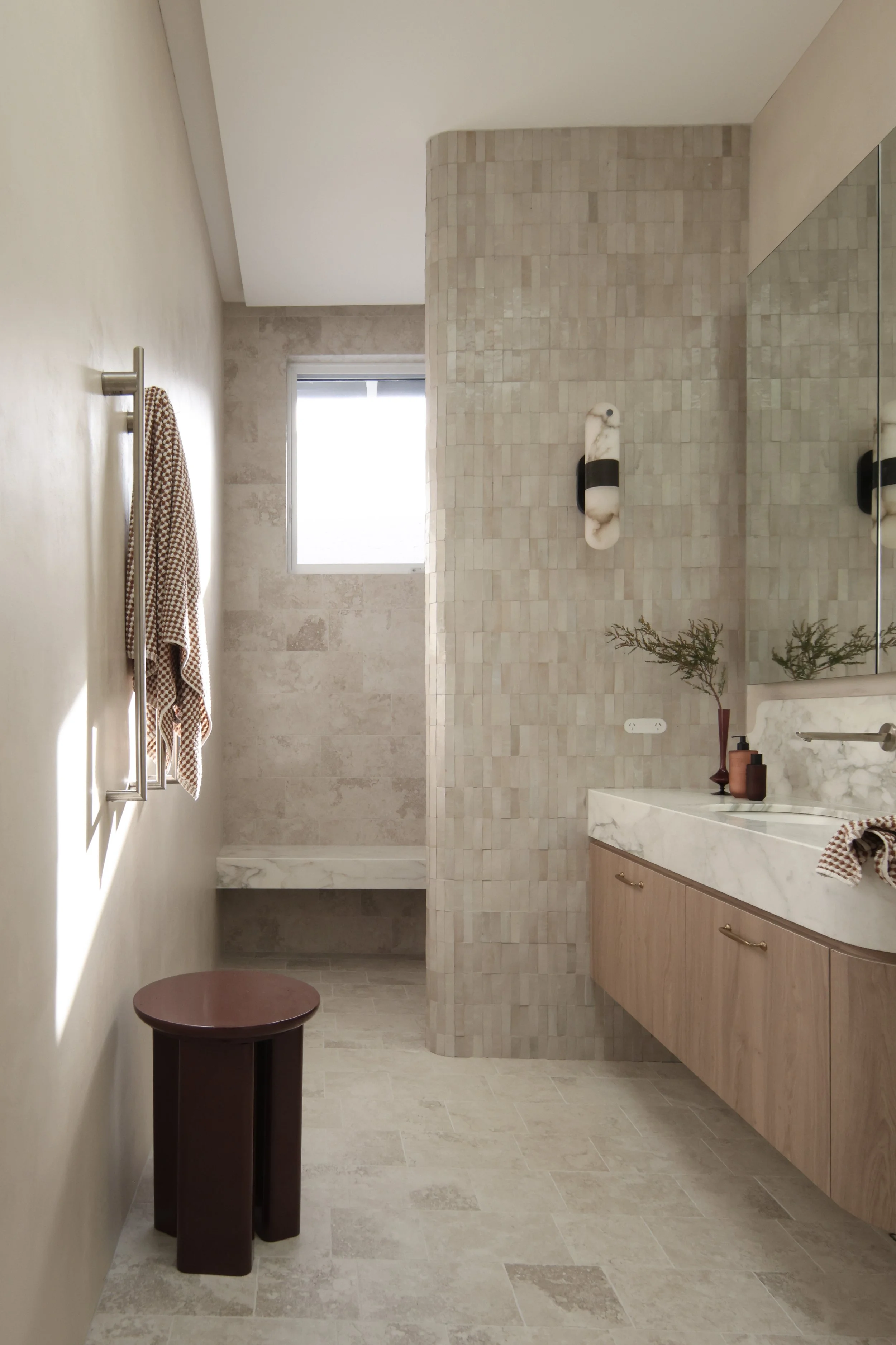 Modern bathroom with hand-cut Moroccan tiles, a marble countertop with a basin, wooden cabinet, potted plant, decorative wall light, a small window, and a towel on a metal towel rack. Light shines through the window onto the wall.