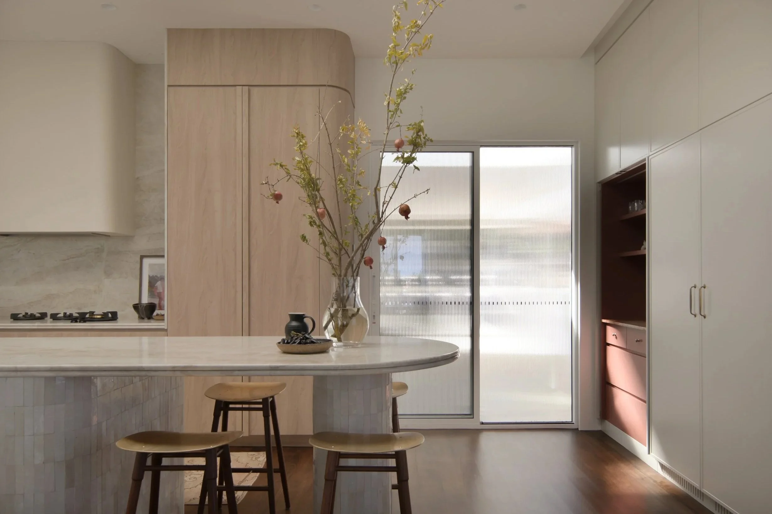 Modern kitchen with a marble island, wooden stools, a vase with branches and pomegranates, a large window, and built-in cabinets in white and terracotta colors.