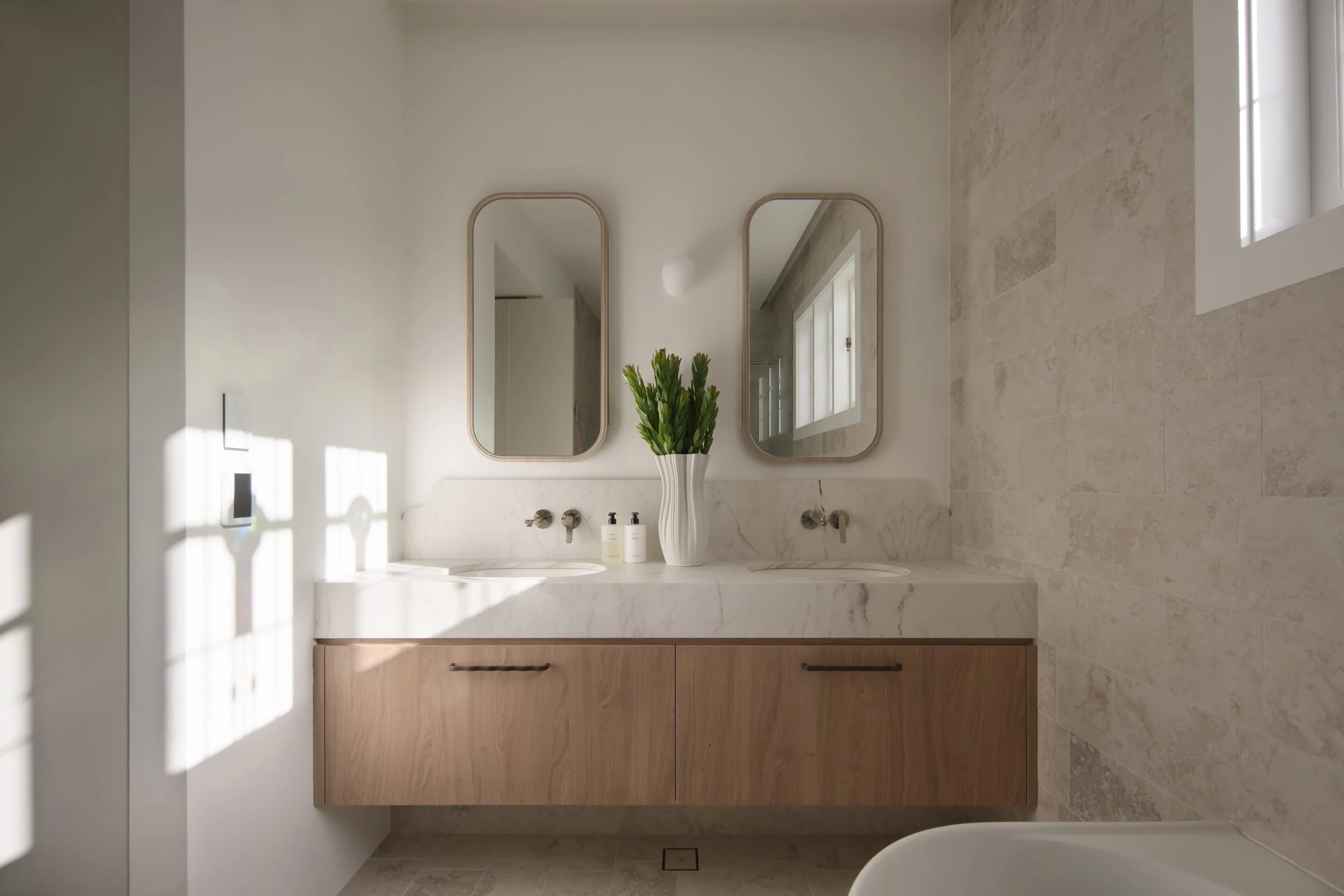 A modern bathroom interior featuring a dual sink marble vanity with a wooden cabinet beneath. Two oval mirrors with light-colored frames hang above the sinks. A tall white vase with green plants is placed on the vanity. The wall is painted white, and a window on the right allows natural light, illuminating beige tiled walls and floors.