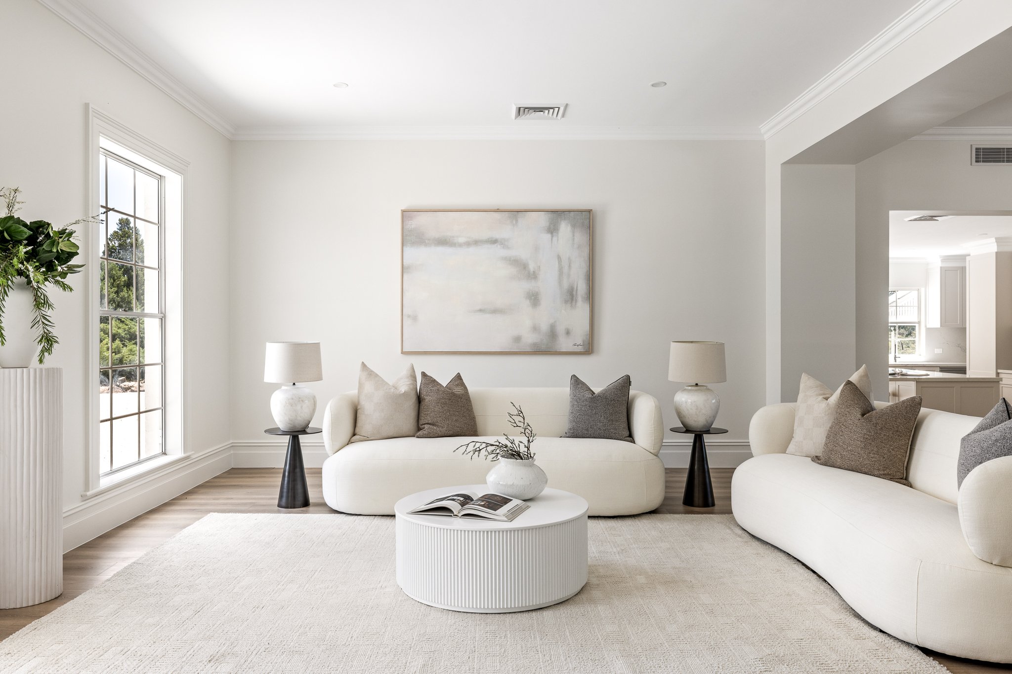 Minimalist living room with two white sofas, neutral-colored pillows, a white round coffee table with a book and a small vase with branches, white walls, a large abstract painting, two table lamps, a window, and a potted plant.