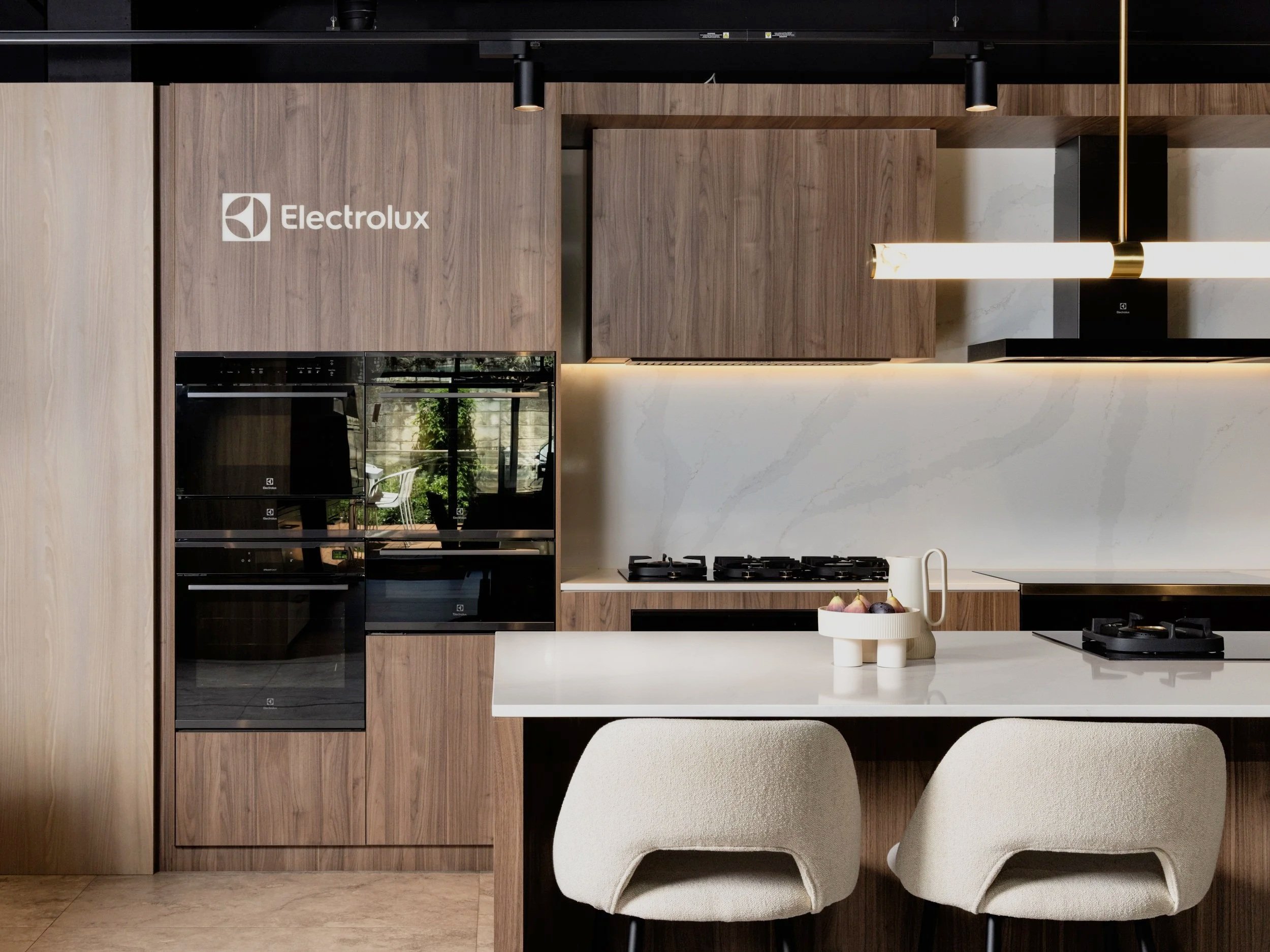 Modern kitchen with wood cabinets, built-in black oven, white countertop, decorative fruit bowl, white pitcher, and beige chairs.