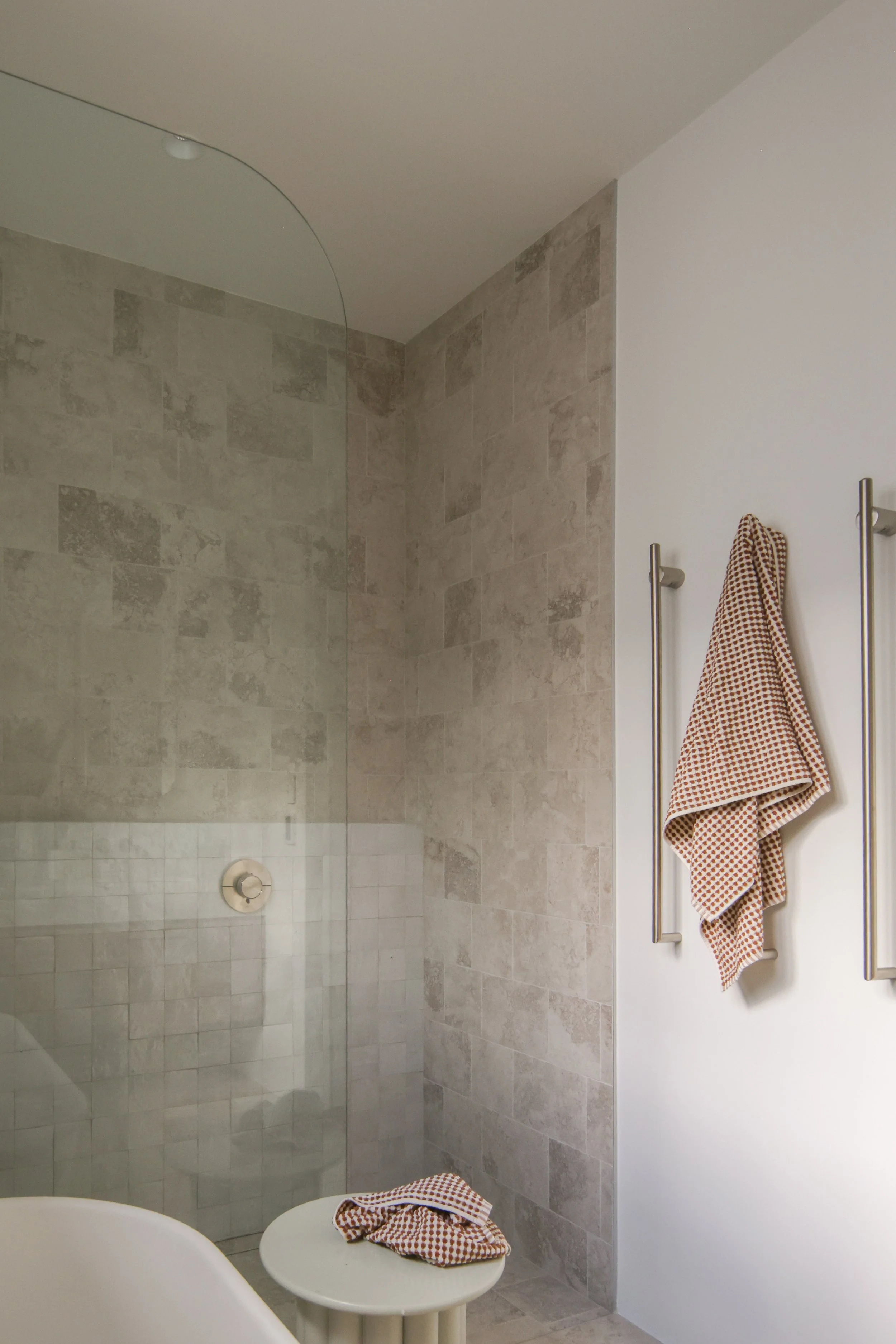 A bathroom with a shower area tiled with beige stone tiles, a glass shower partition, a white wall with two towel bars, and a checkered towel hanging on one of the bars. A small round table with a matching checkered cloth and a chair are at the foreground.