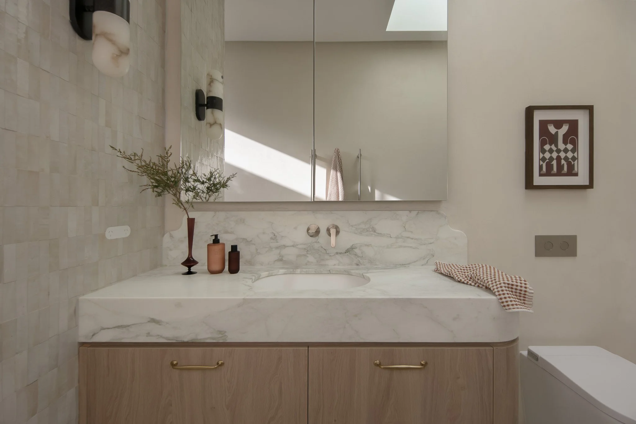 Bathroom mirror reflecting a vanity with a marble countertop, a framed picture on the wall, three decorative vases with a plant, and a neatly folded cloth on the counter.