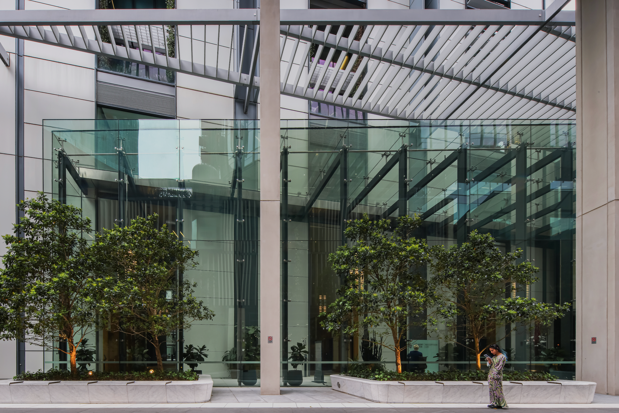 Modern building with large glass windows, green trees in planters, and a person standing outside looking at their phone.