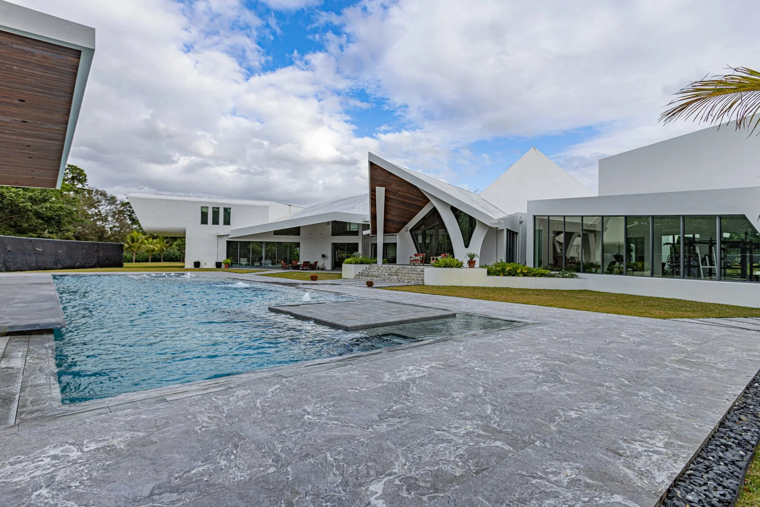 A wide-angle view of the pool and natural stone patio deck, featuring a raised spa or sun shelf, lush green surroundings, and the home's bold architectural silhouette in the background.