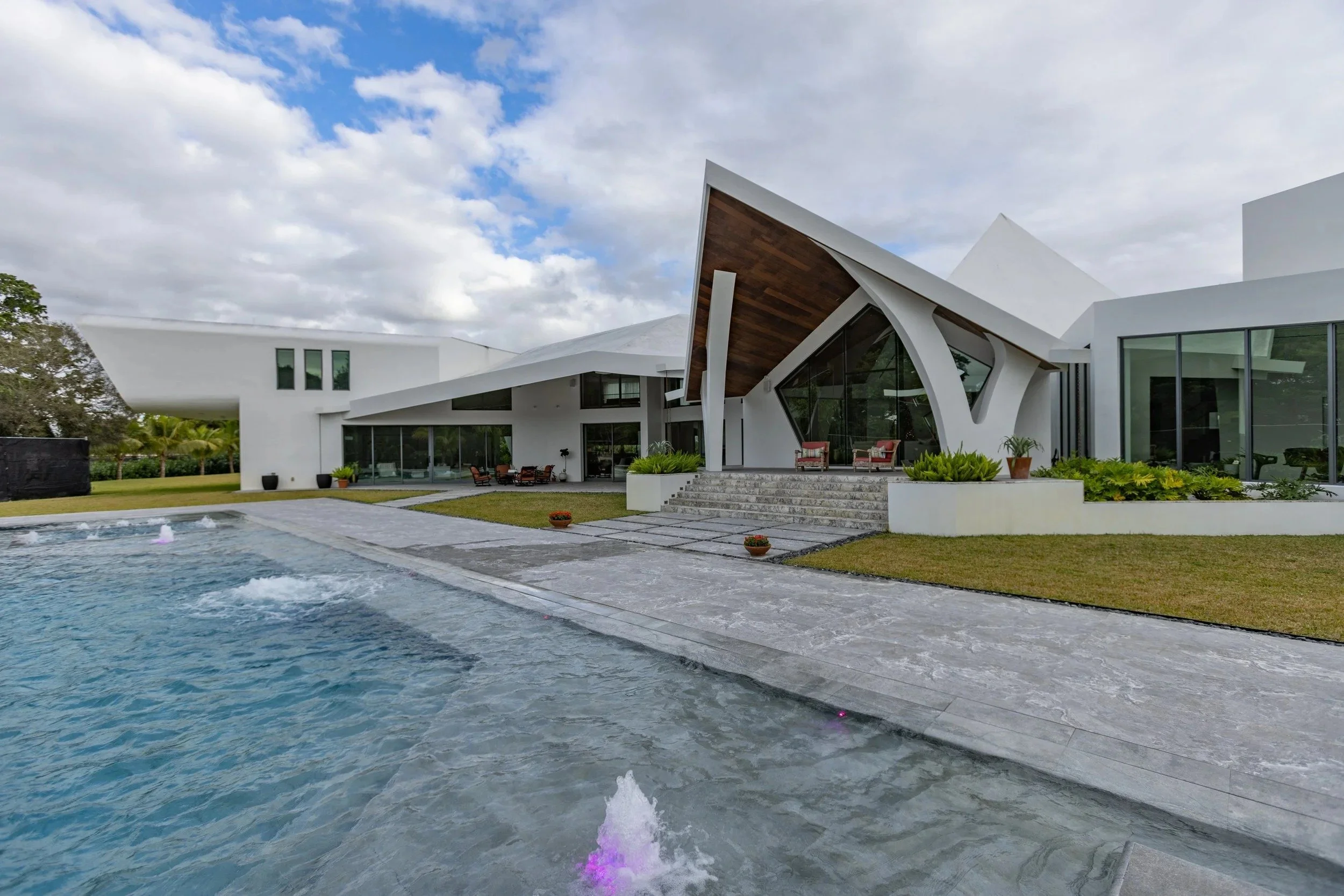 Ground-level view of the pool and home exterior showing the expansive stone pool deck, unique asymmetrical roofline, and an active deck jet — a stunning example of our luxury residential pool work.