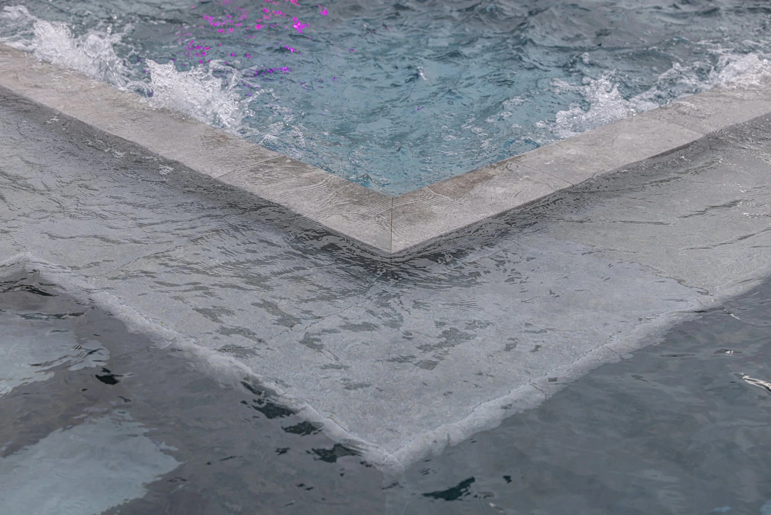 A detail close-up of the pool corner featuring precision-cut mitered stone coping with active water splashing — a hallmark of our meticulous craftsmanship and high-quality finish work.
