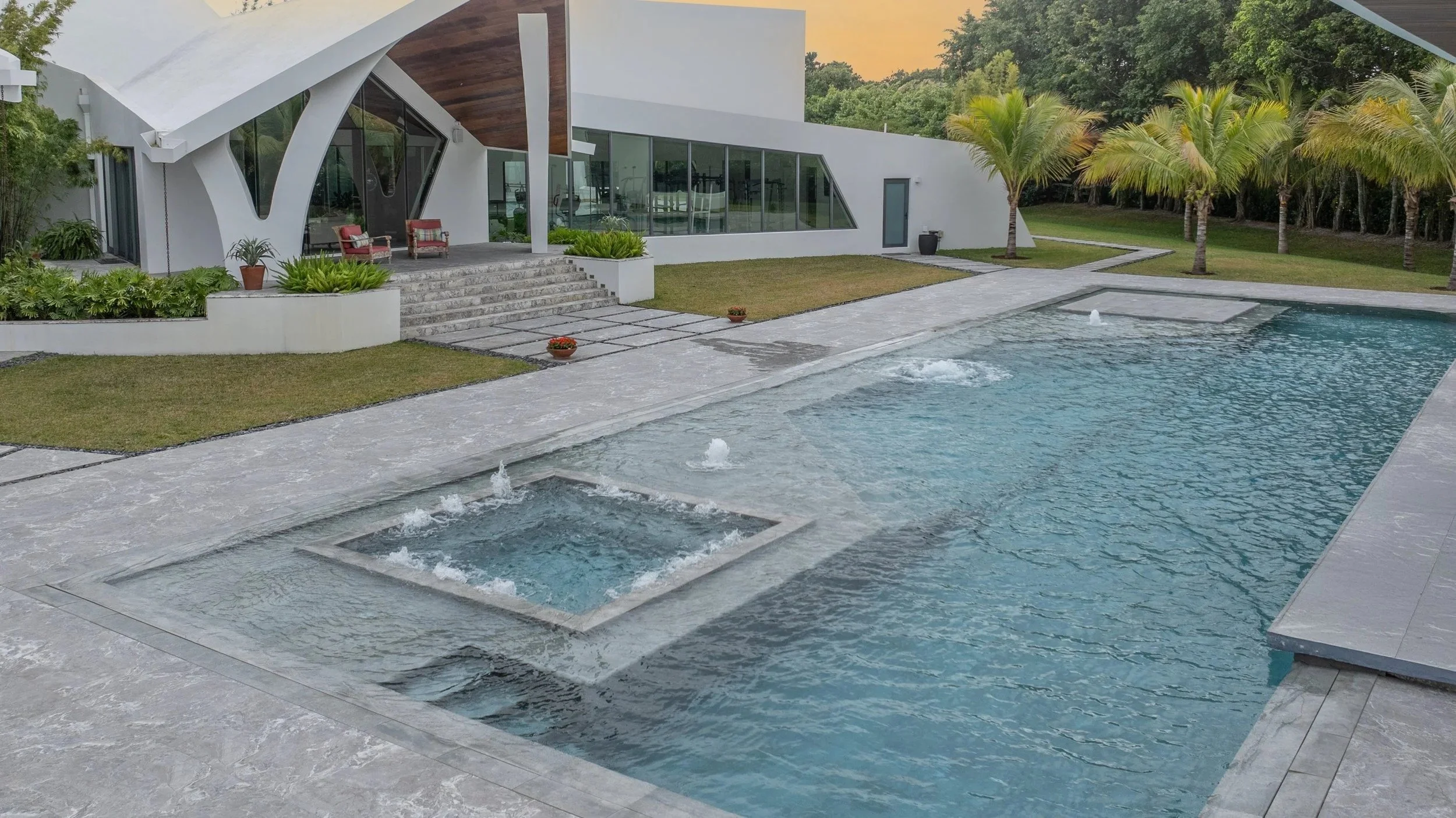 Golden hour poolside view showcasing an integrated square spa with bubblers alongside the main pool, lush tropical palms, and the home's iconic roofline — the perfect outdoor living retreat.