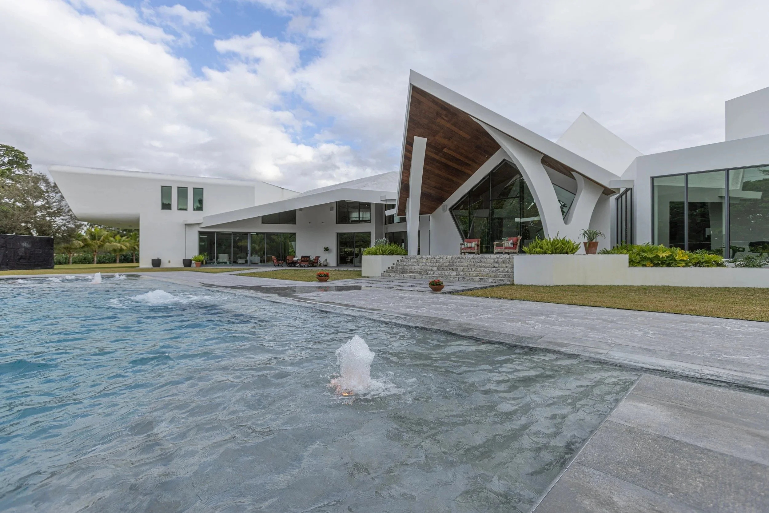 A poolside view featuring an active deck jet fountain set against a stunning modern home with dramatic angular architecture — showcasing the full resort-style experience our pool construction delivers.
