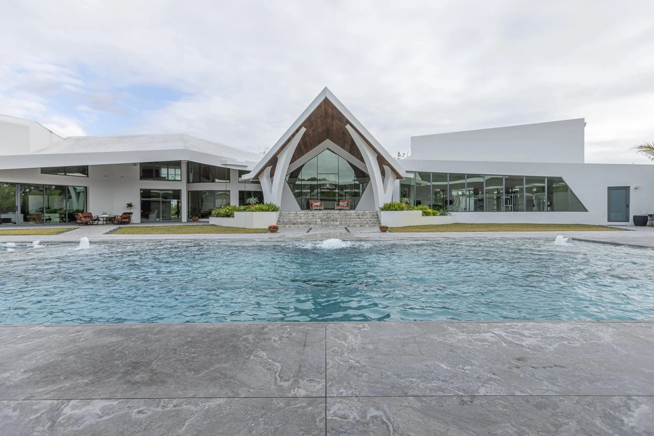 A large luxury pool perfectly framed by the home's striking architectural roofline, featuring crystal-clear water, deck jet bubblers, and premium stone coping — a seamless blend of elegant design and expert pool construction.