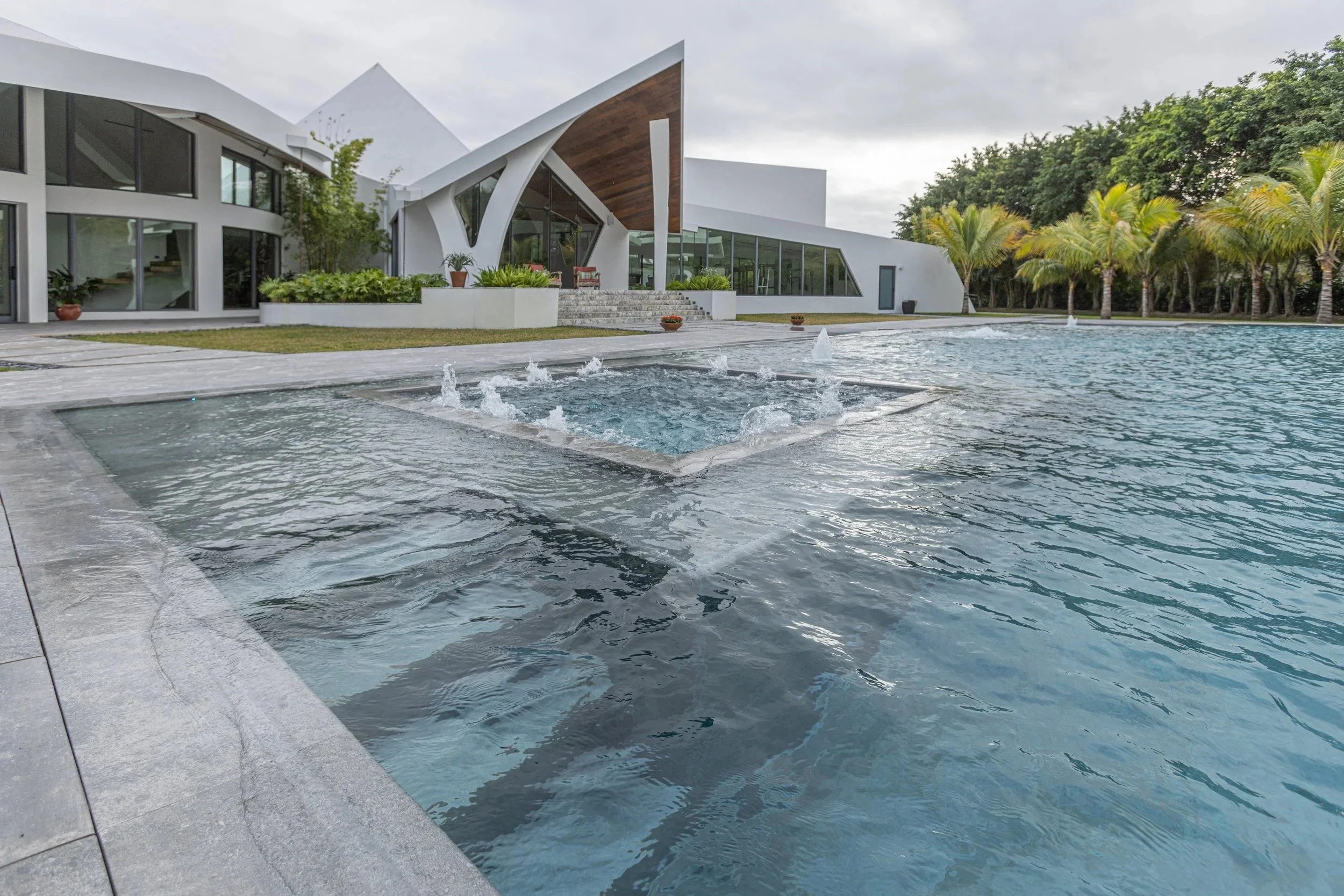 The integrated square spa fountain overflows into the main pool, set against the home's iconic architectural silhouette and lush palm tree backdrop — an eye-catching water feature centerpiece.
