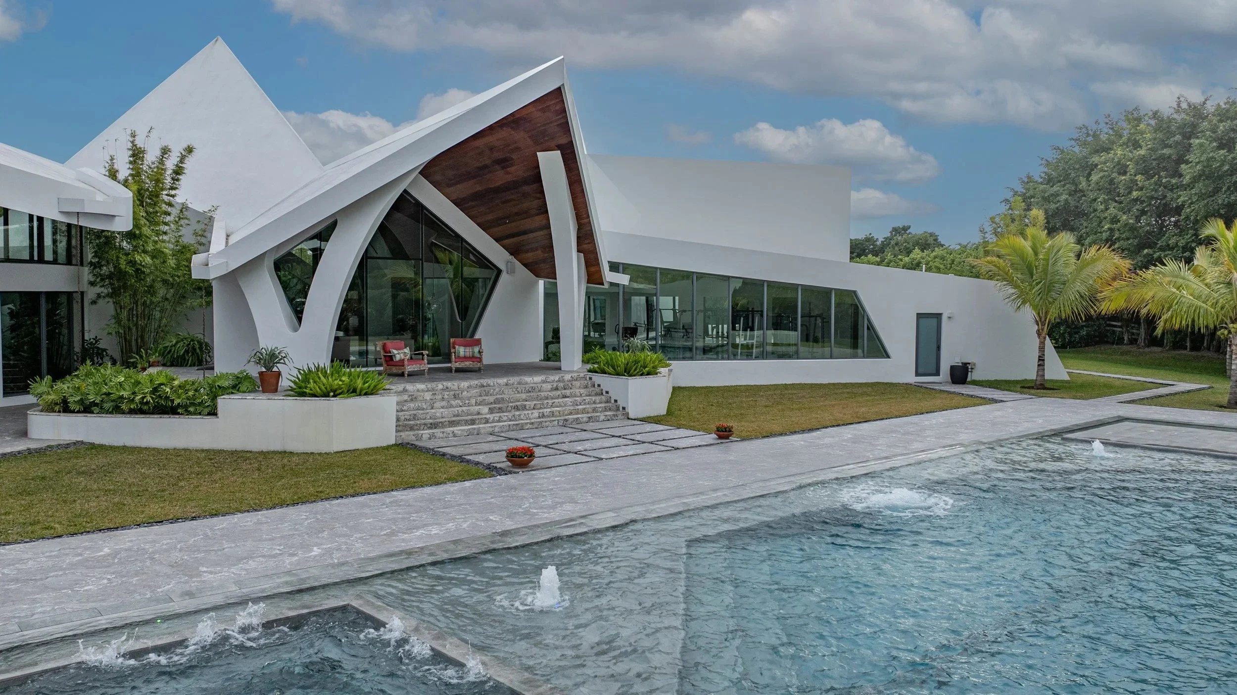 A crisp blue-sky view of the pool with active deck jet fountains, framing the home's bold angular architecture and wood-accented entryway — a project where every detail reflects quality and vision.