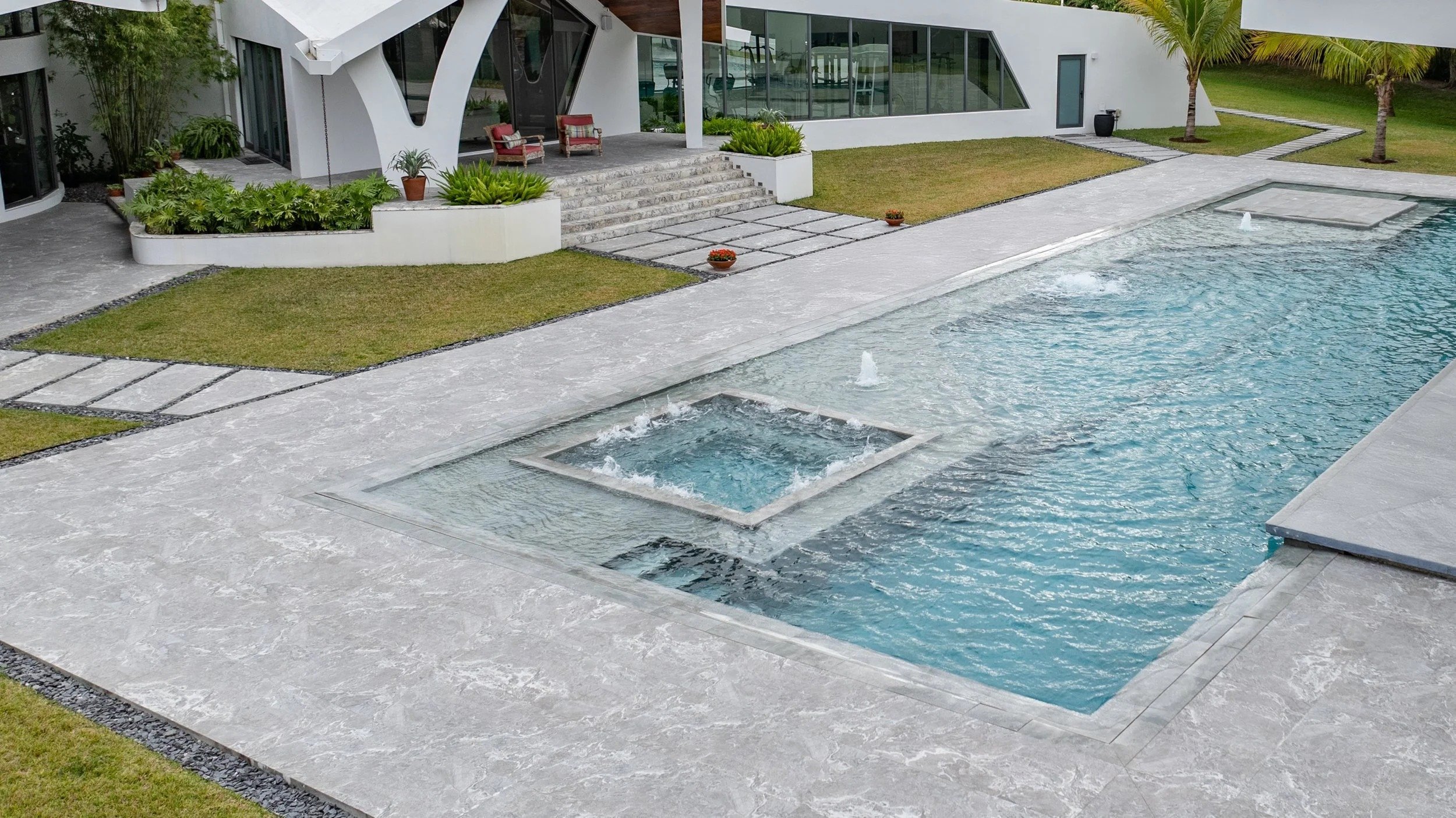 Daytime view of the pool and integrated spa combination set within a premium stone deck, with the home's dramatic arched entryway and curated garden landscaping creating an inviting outdoor oasis.