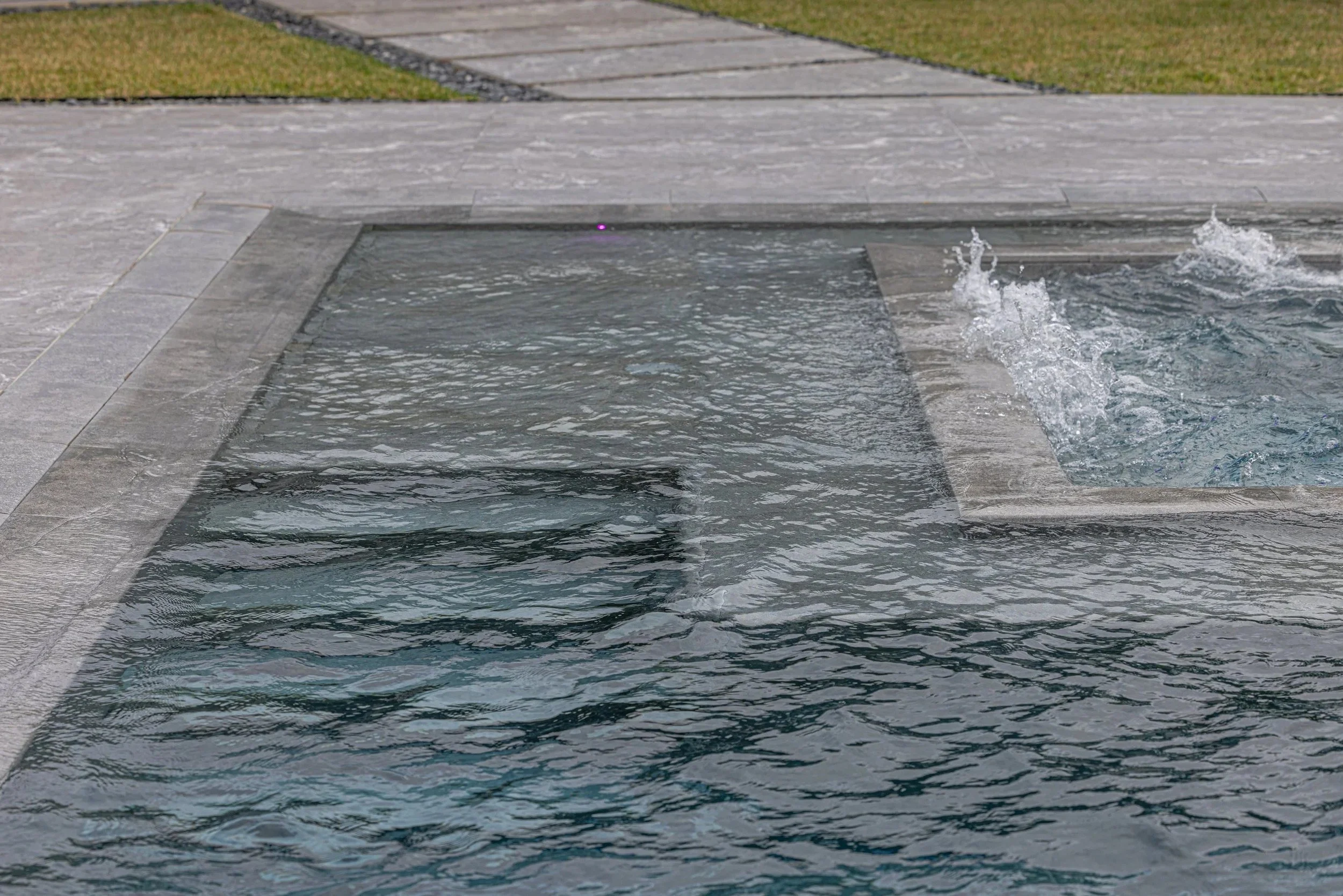 A water-level close-up capturing the dynamic movement of a pool bubbler jet, highlighting the crisp stone coping detail and the quality of water features we incorporate into every build.