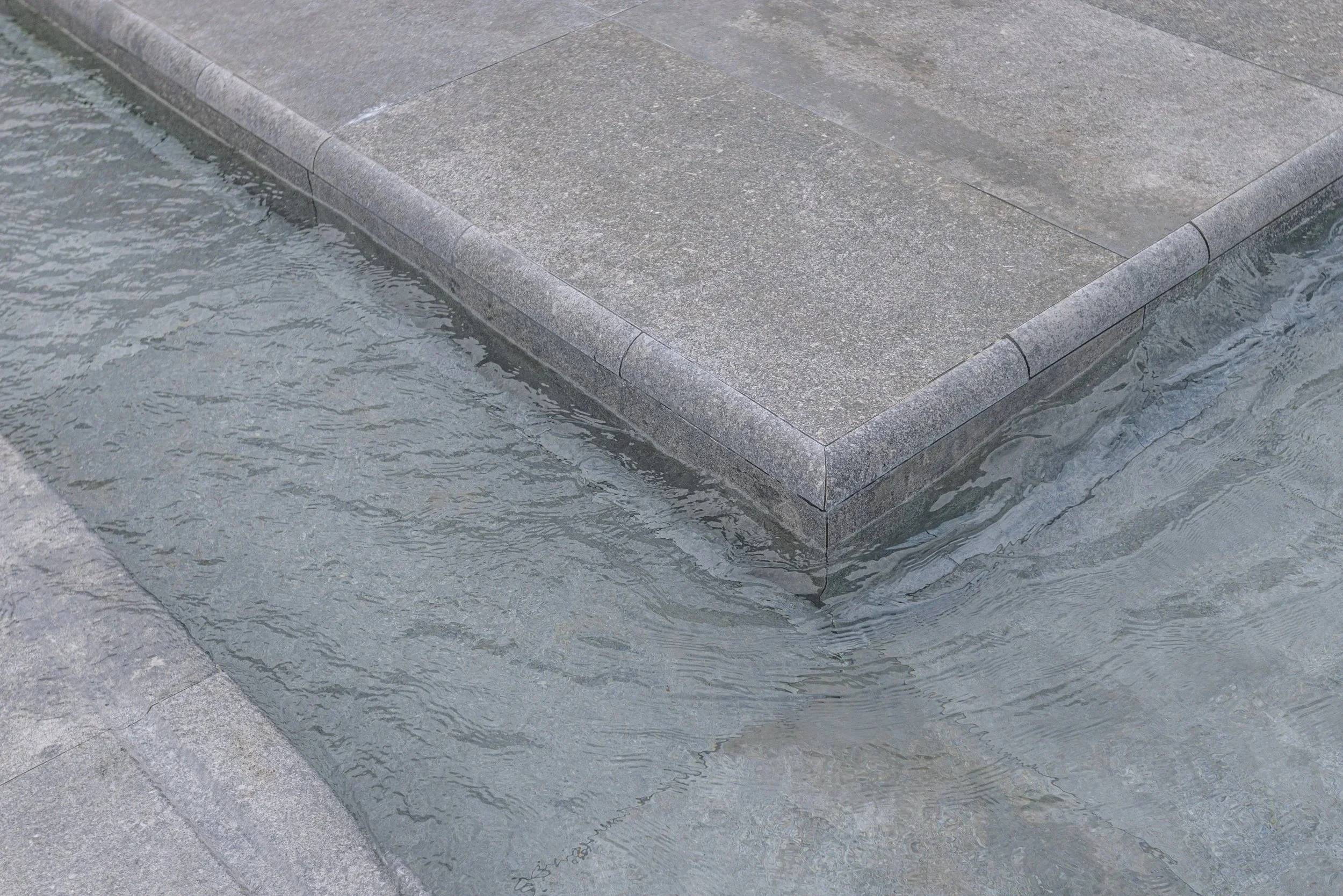 Close-up of the pool's raised tanning ledge edge, showcasing the precision bullnose stone coping installation and water flowing cleanly around the structure — quality craftsmanship visible in every inch.