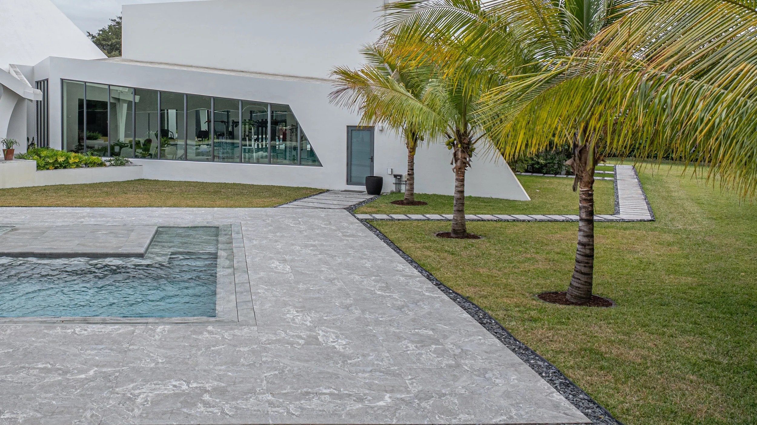 Poolside view framed by swaying palm trees, showcasing the pool's overflow edge, premium stone deck, and the home's dramatic glass facade — a resort-quality outdoor space built to impress.