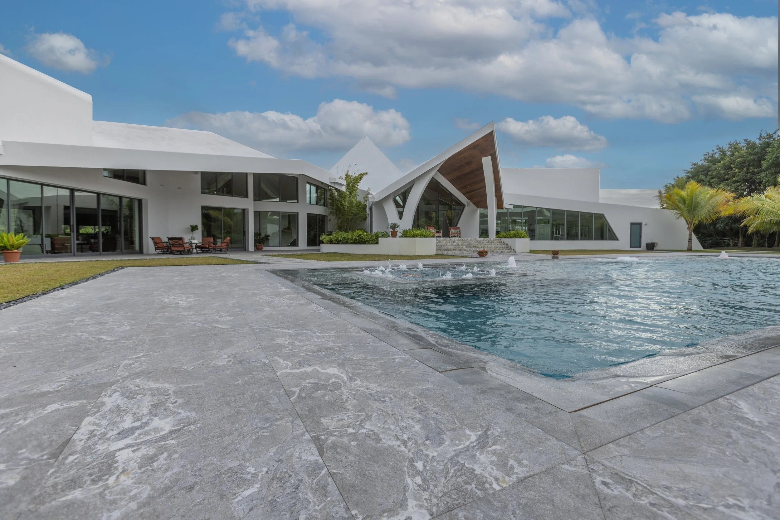 Sun-drenched view of the natural stone pool deck leading to the pool and a sheer descent waterfall wall, with the home's stunning angular silhouette set against a bright blue sky.