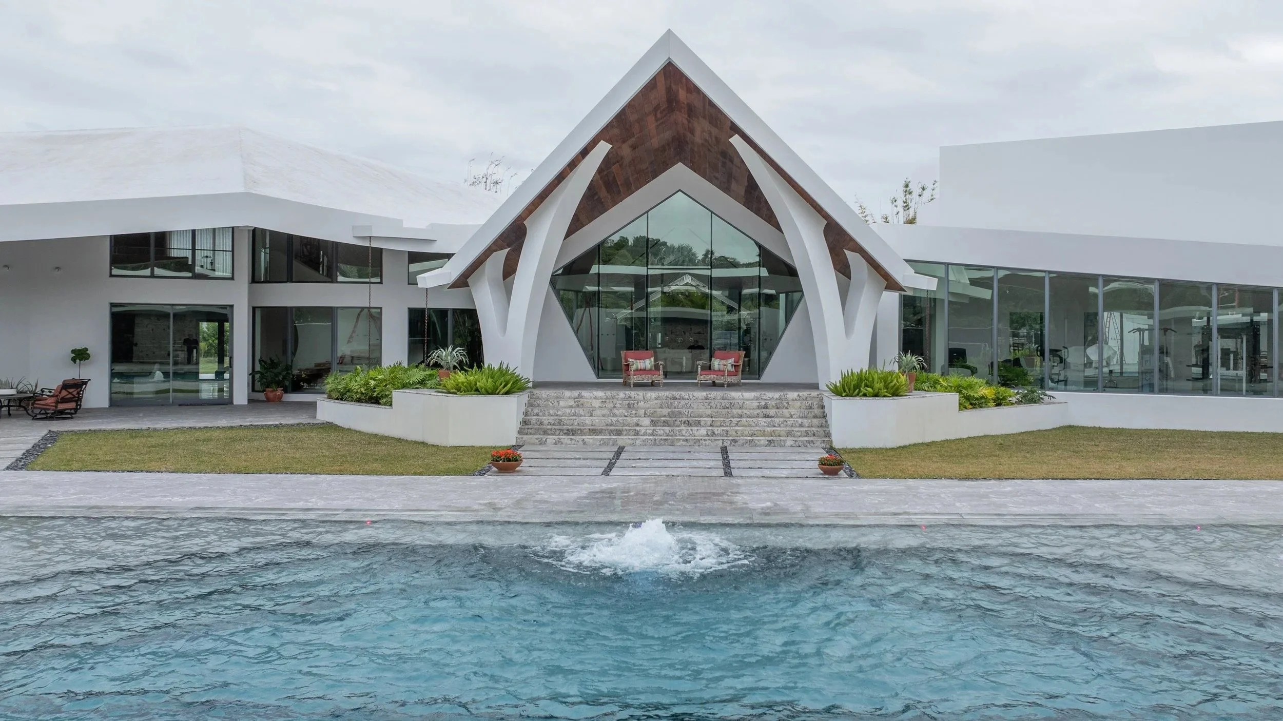 A symmetrical pool-to-home view showcasing the striking A-frame glass entry, stone staircase, and centered deck jet bubbler — where architectural excellence meets masterful pool design.