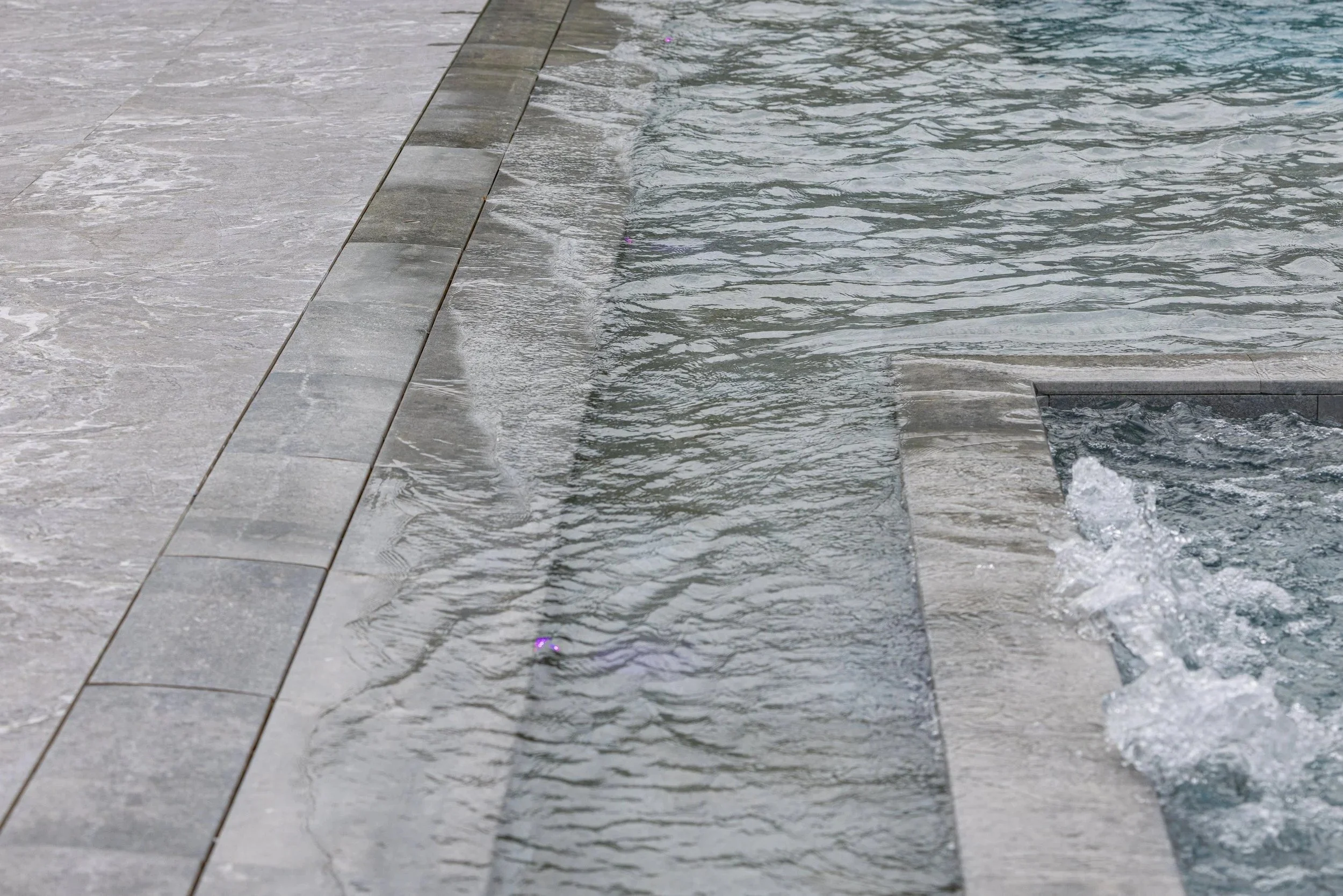 A close-up detail of a custom pool overflow edge, highlighting the precision stone coping and the smooth water sheet cascading over the spillway — a signature finish in our high-end pool builds.
