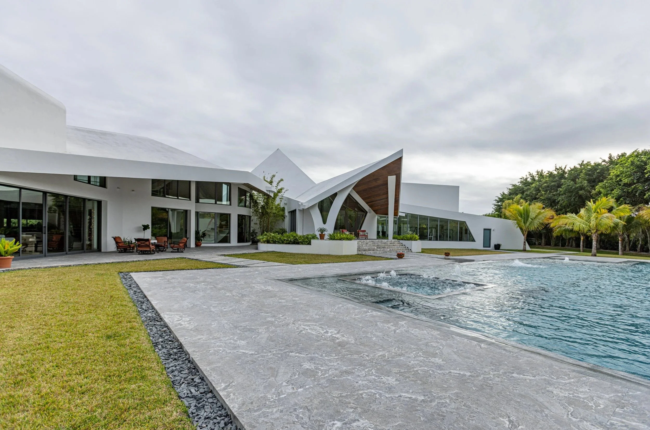 A full property view capturing the pool, expansive stone deck, manicured lawn, and the home's sweeping modern architecture surrounded by tropical landscaping — the complete luxury outdoor environment we build.