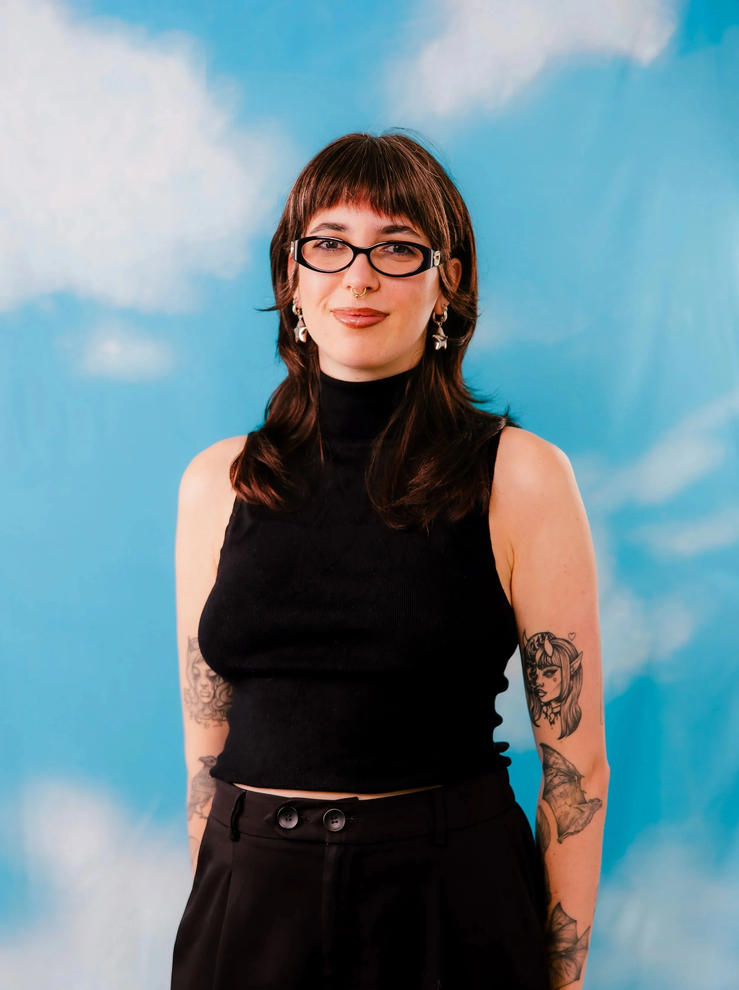 A young woman with brown hair, glasses, and tattoos on her arms, standing against a sky with clouds painted on the background.