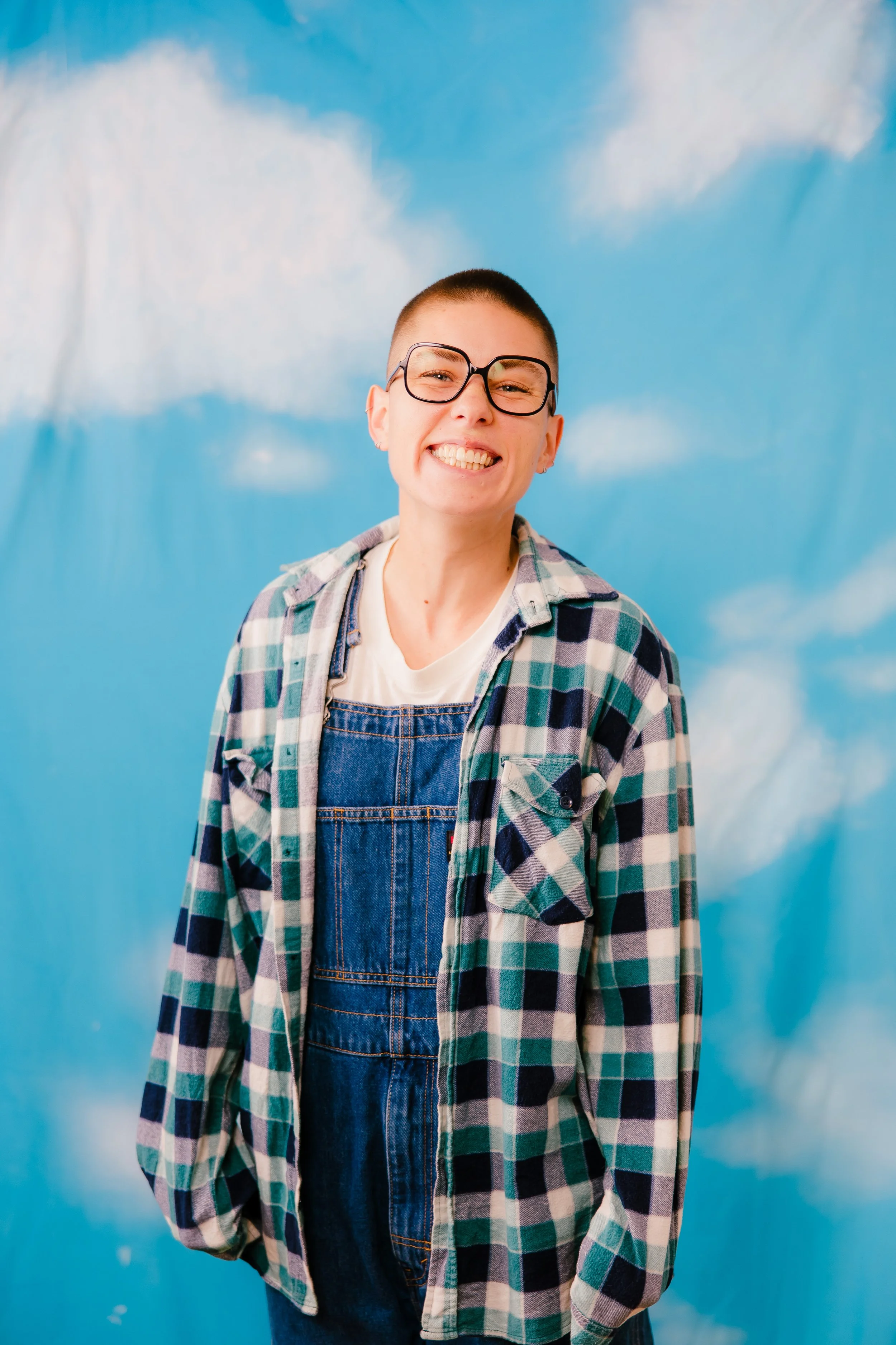 A person with short hair, glasses, and earrings smiling, wearing a plaid shirt over denim overalls. Standing against a sky with clouds painted on the background.