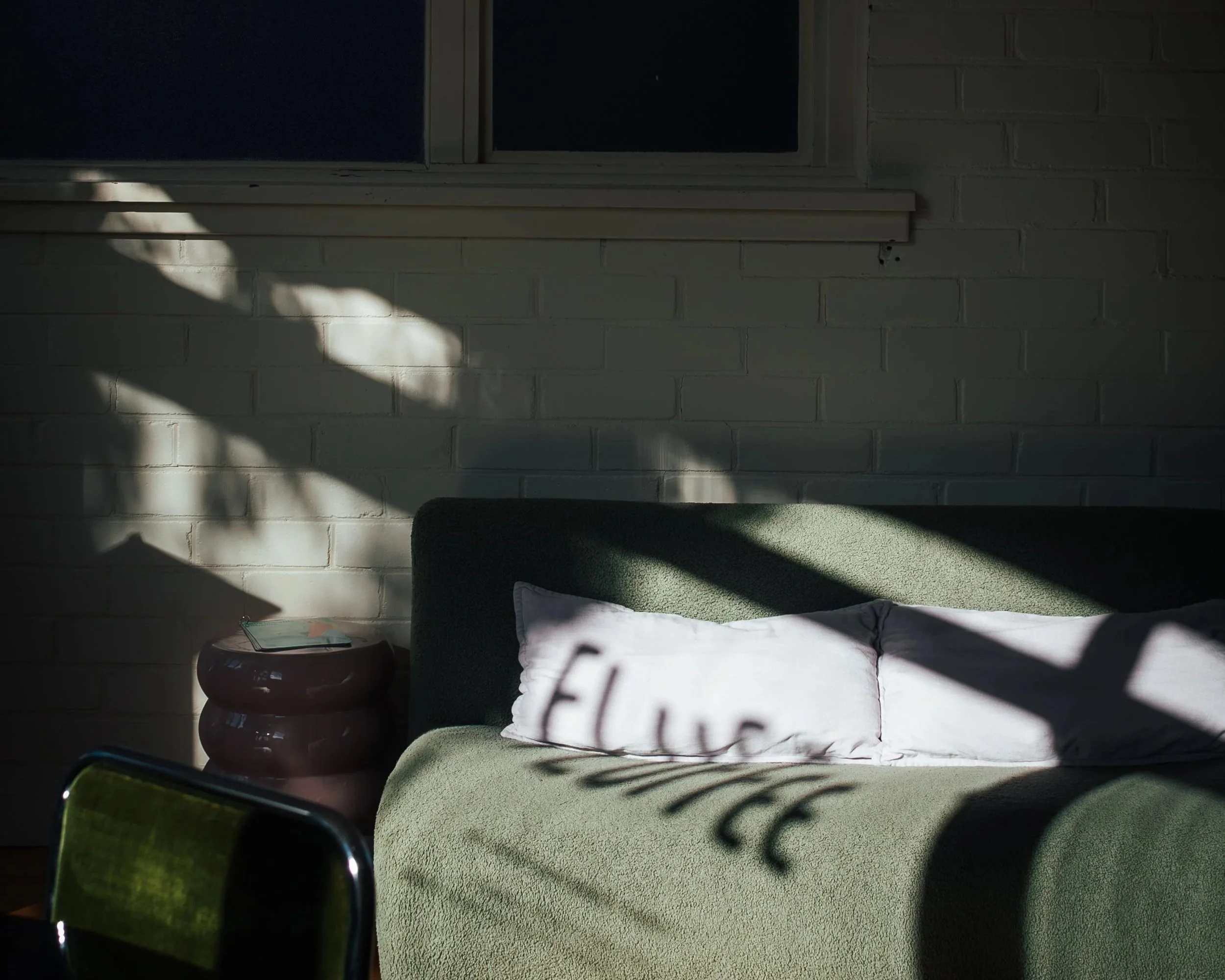 A cozy indoor scene with a bright, white brick wall illuminated by sunlight. There is a green upholstered chair with a white pillow. Next to the chair is a small, round, stacked ceramic table.
