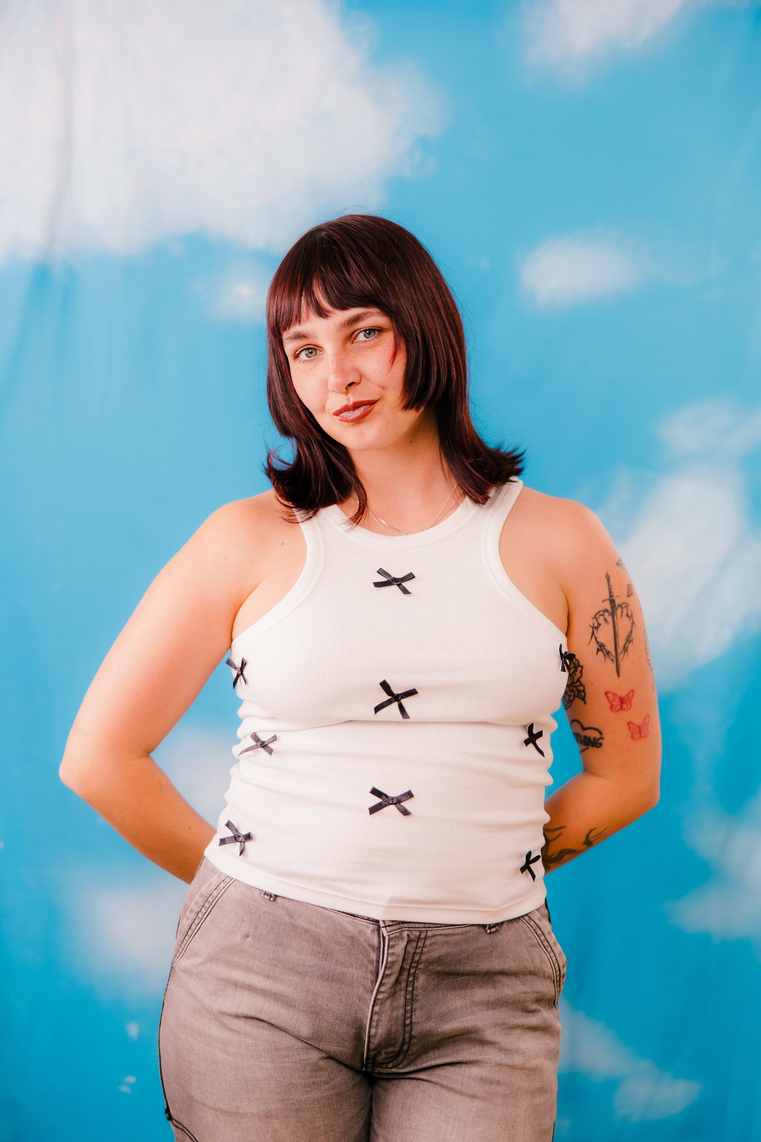 A woman with short dark hair and multiple tattoos on her arms, wearing a white tank top with small black bows and gray pants, standing against a sky with clouds painted on the background.